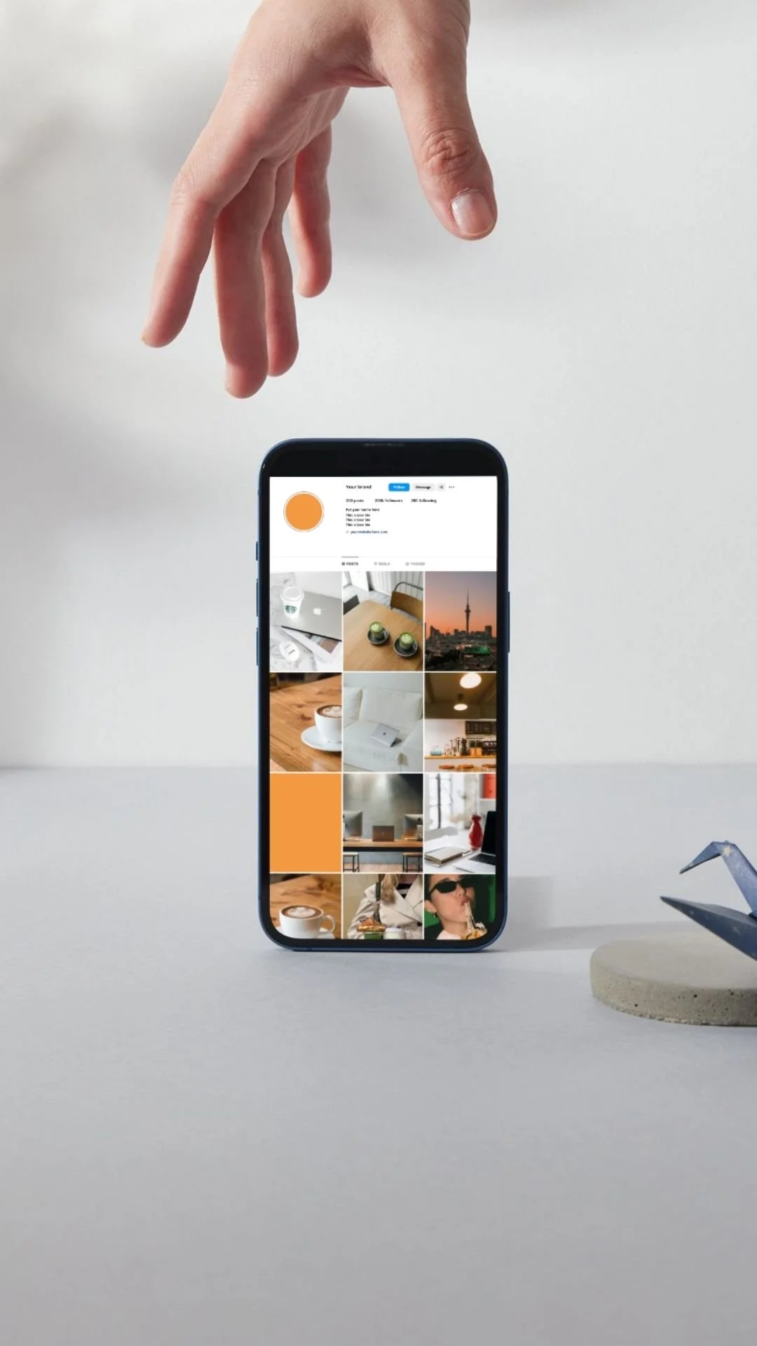 A hand is hovering above a smartphone displaying a photo gallery app on a plain white surface with a gray bobble-head bird figurine on the right.
