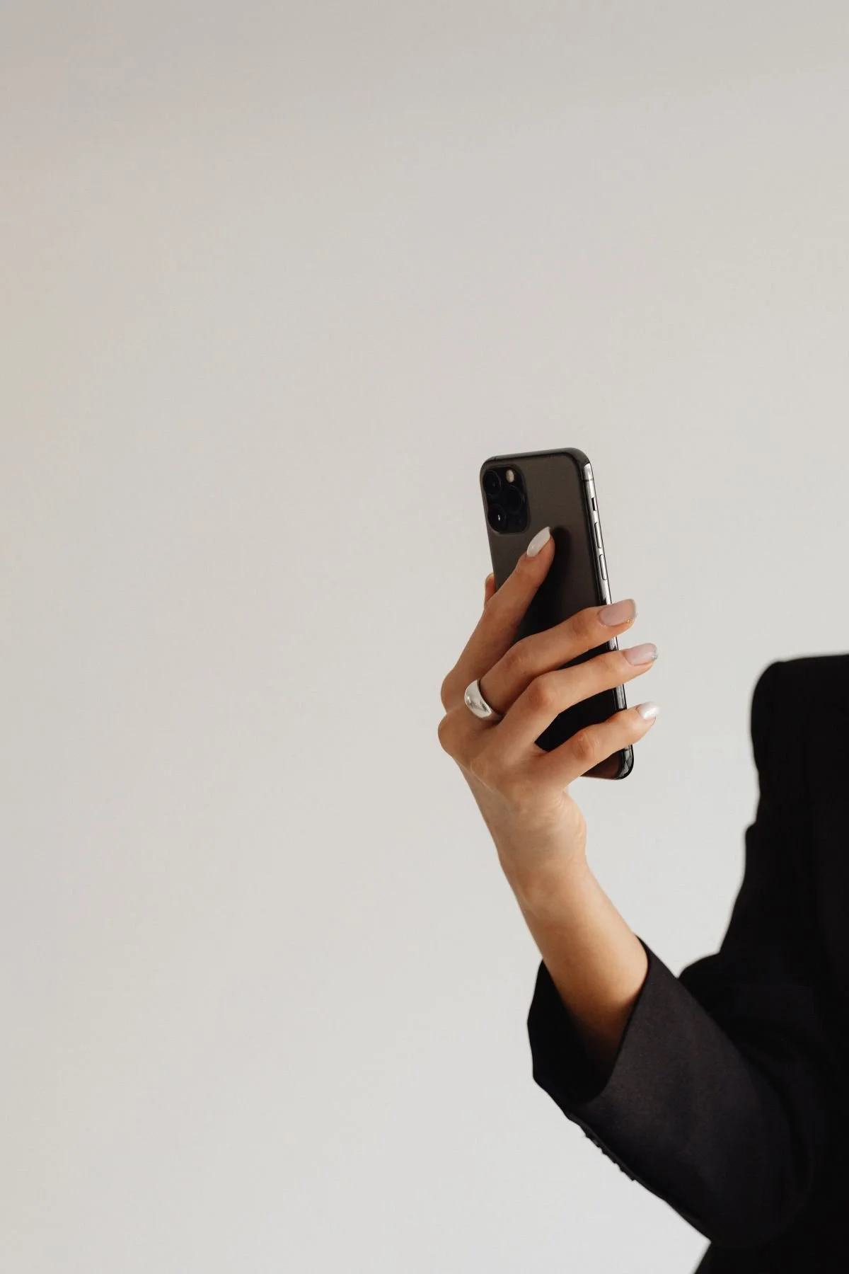 Person holding a smartphone with a black case and a silver ring on their finger, wearing a black blazer, against a plain white background.