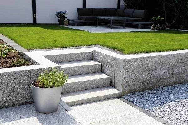 Garten mit grüner Rasenfläche, grauen Treppen und Pflanzkübel, Sitzbereich mit schwarzem Sofa im Hintergrund.