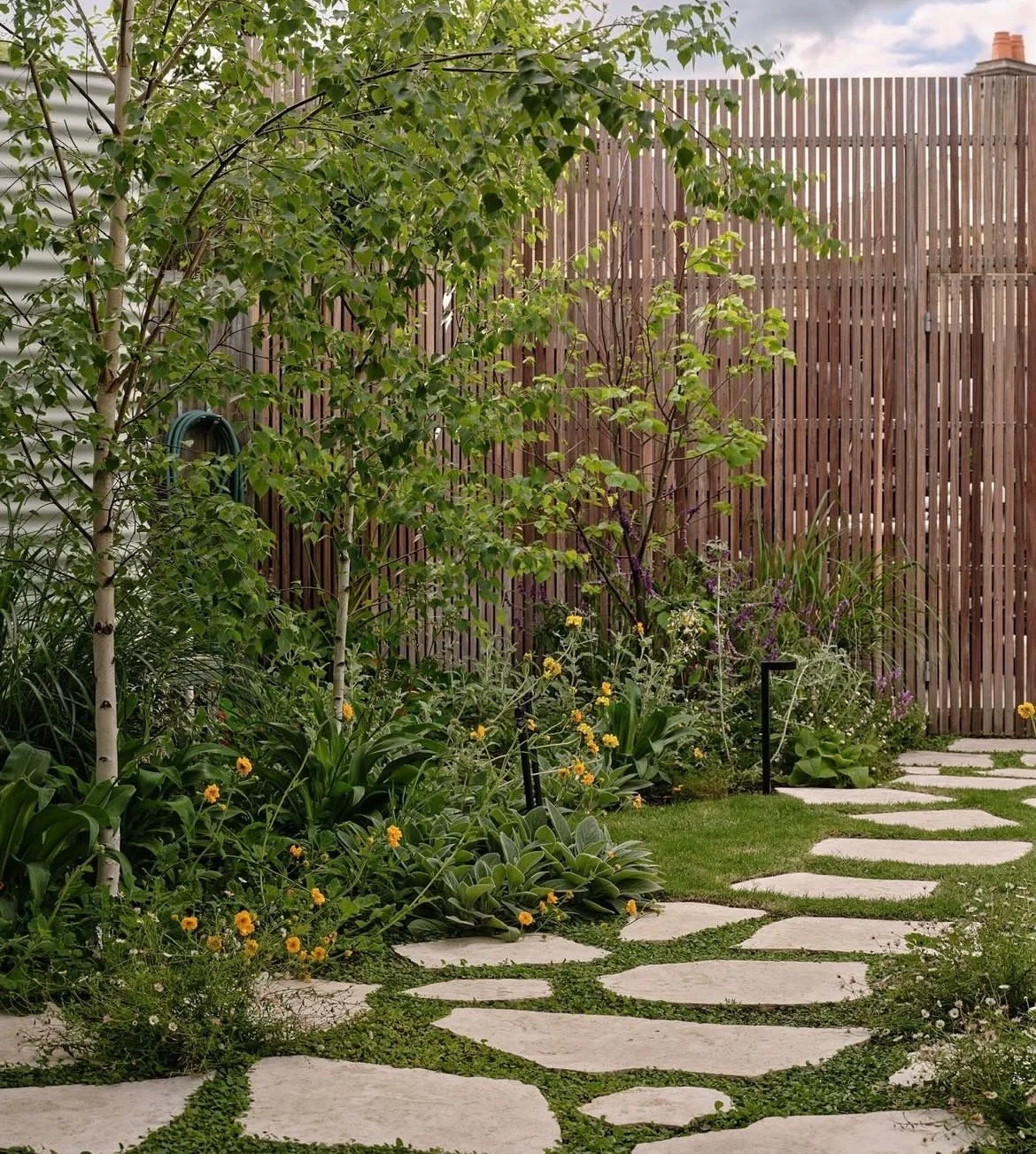 Garten mit Steinstraße, Bäume, Blumen und Holzzaun im Hintergrund.
