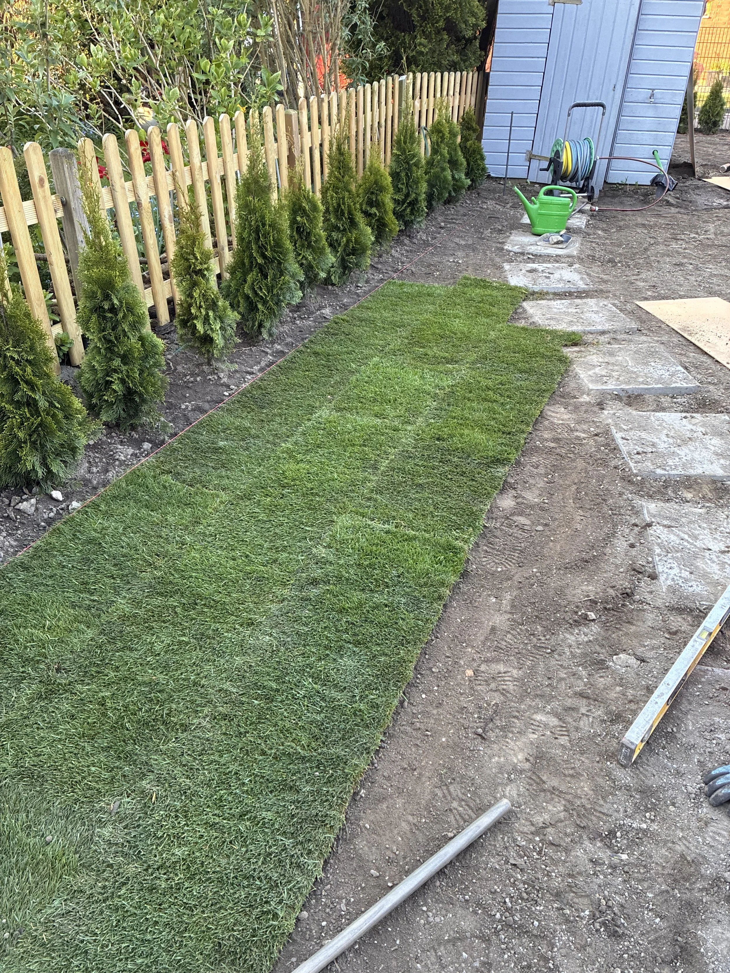 Der Garten wird vorbereitet, mit einem sauberen Rasen und einem Weg aus Pflastersteinen, die auf dem Boden verlegt werden. Es gibt Werkzeug und Gartenschläuche zum Gießen.