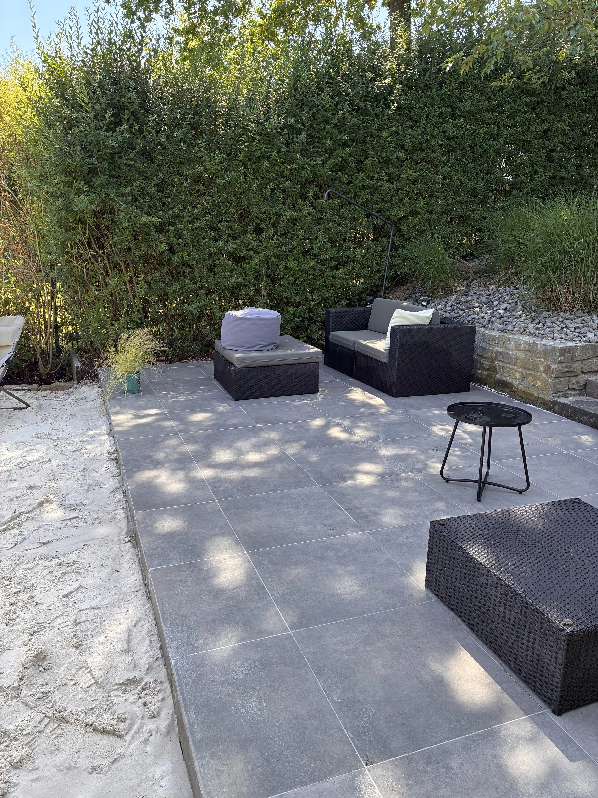 Gartenmöbel mit dunklem Rattan und grauen Kissen auf einer Terrassenfläche, umgeben von starkem Bewuchs und einem Gemüsegarten mit Steinbeeten.