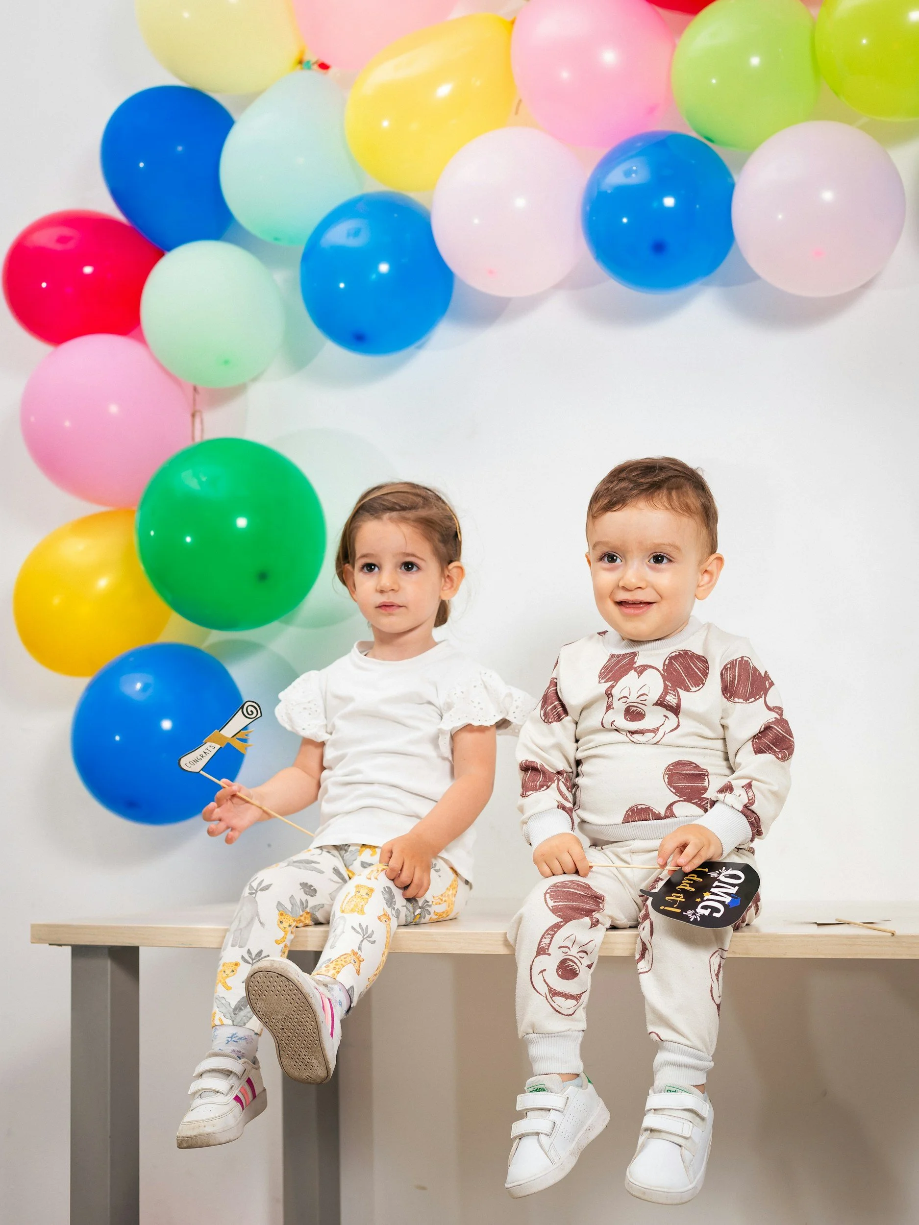 Two young children sit on a wooden bench in front of a white wall decorated with colorful balloons, one girl holding a small paper sign and a boy holding a birthday card, both wearing casual clothing and white sneakers.