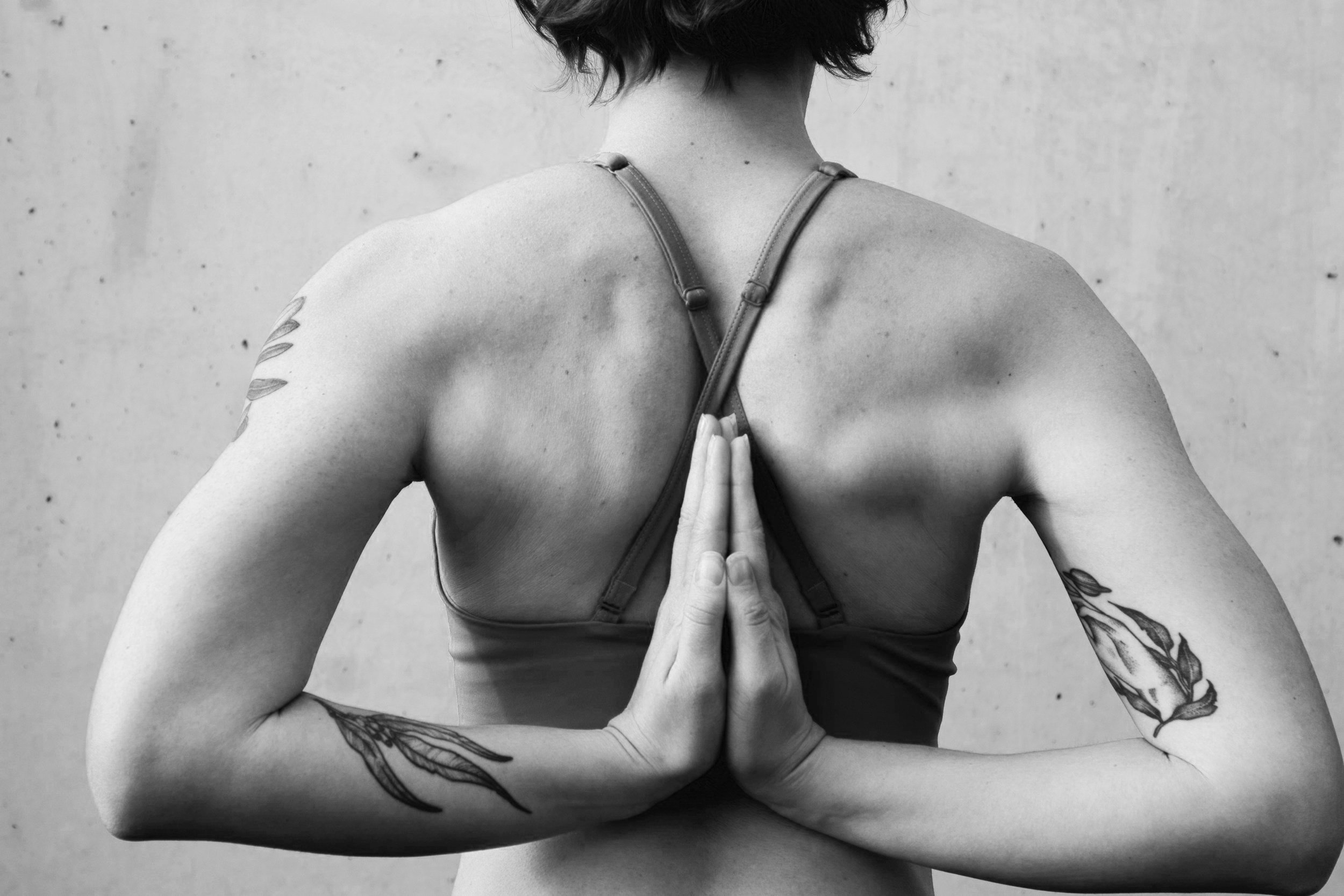 En kvinna med tatueringar på armarna och en liten tatuering på axeln, hon är i yogaposition med händerna i bönläge framför ryggen, fotograferad i svartvitt mot en enkel vägg.