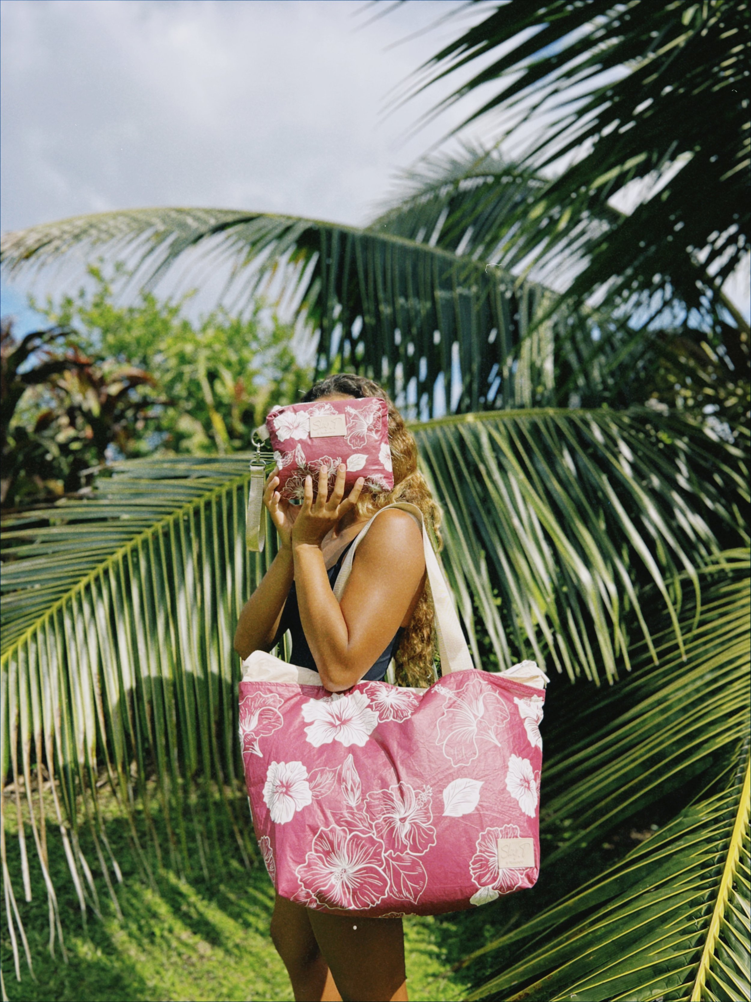 Femme portant un sac à main et un sac de plage à motifs floraux roses et blancs, se tenant devant de grandes feuilles de palmier dans un environnement tropical