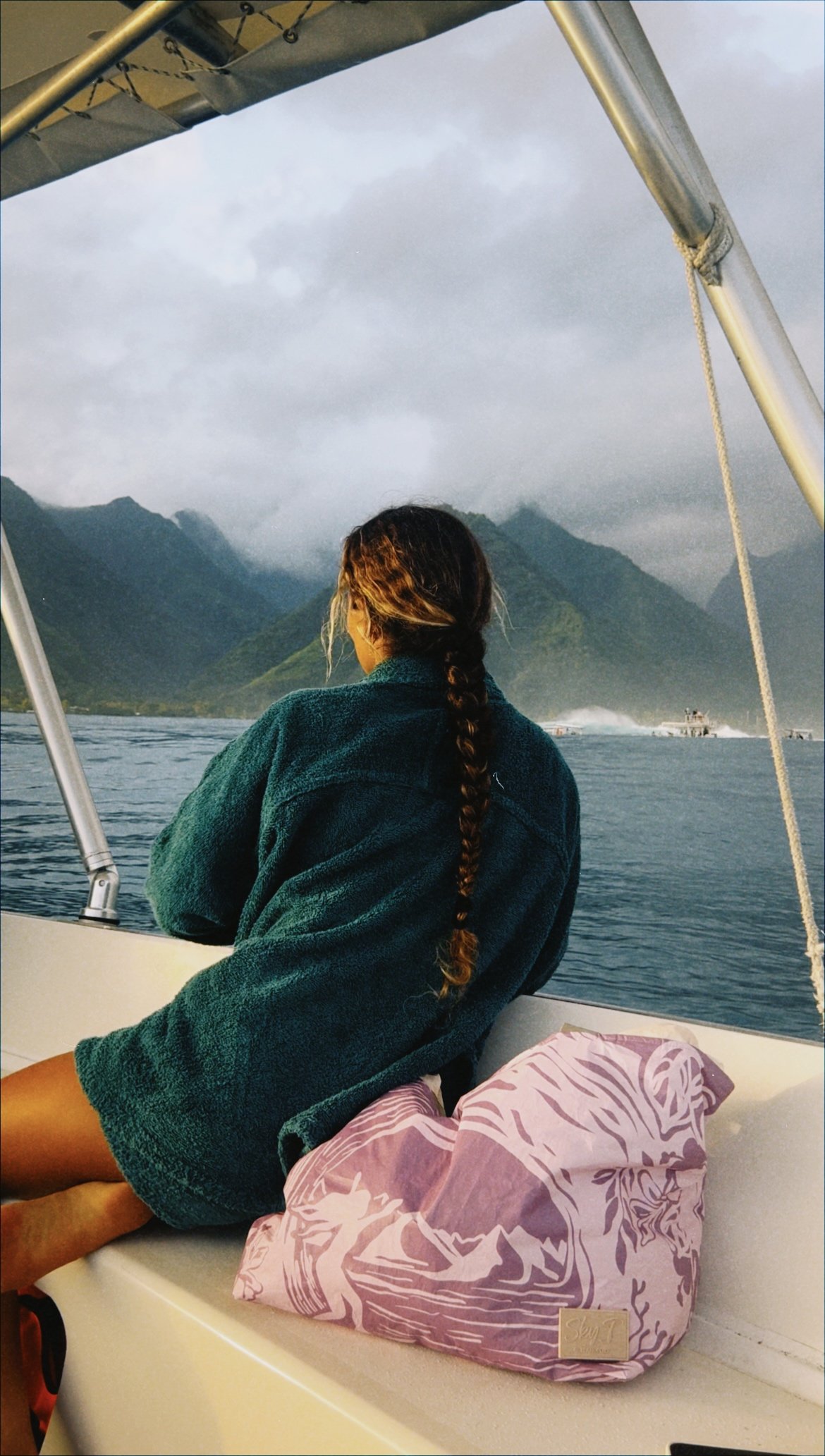 Femme assise sur un bateau regardant vers des montagnes et un lac, portant un pull vert foncé, avec un sac en tissu rose avec un motif de tigre