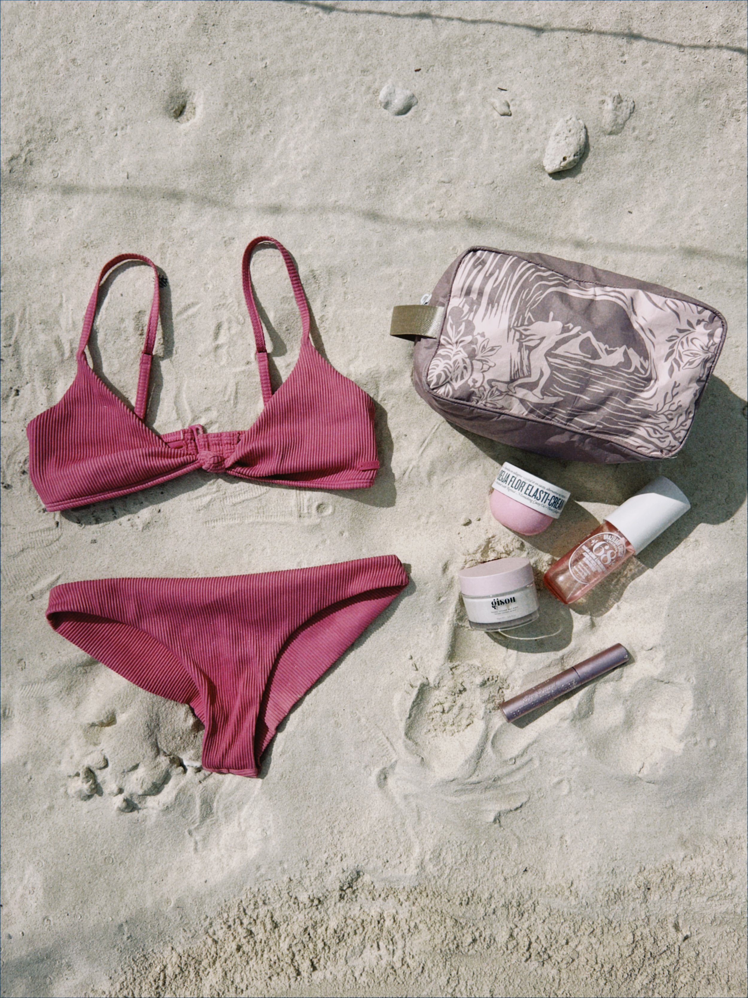 Une tenue de bikini rose avec des produits de protection solaire et un sac de plage sur le sable.