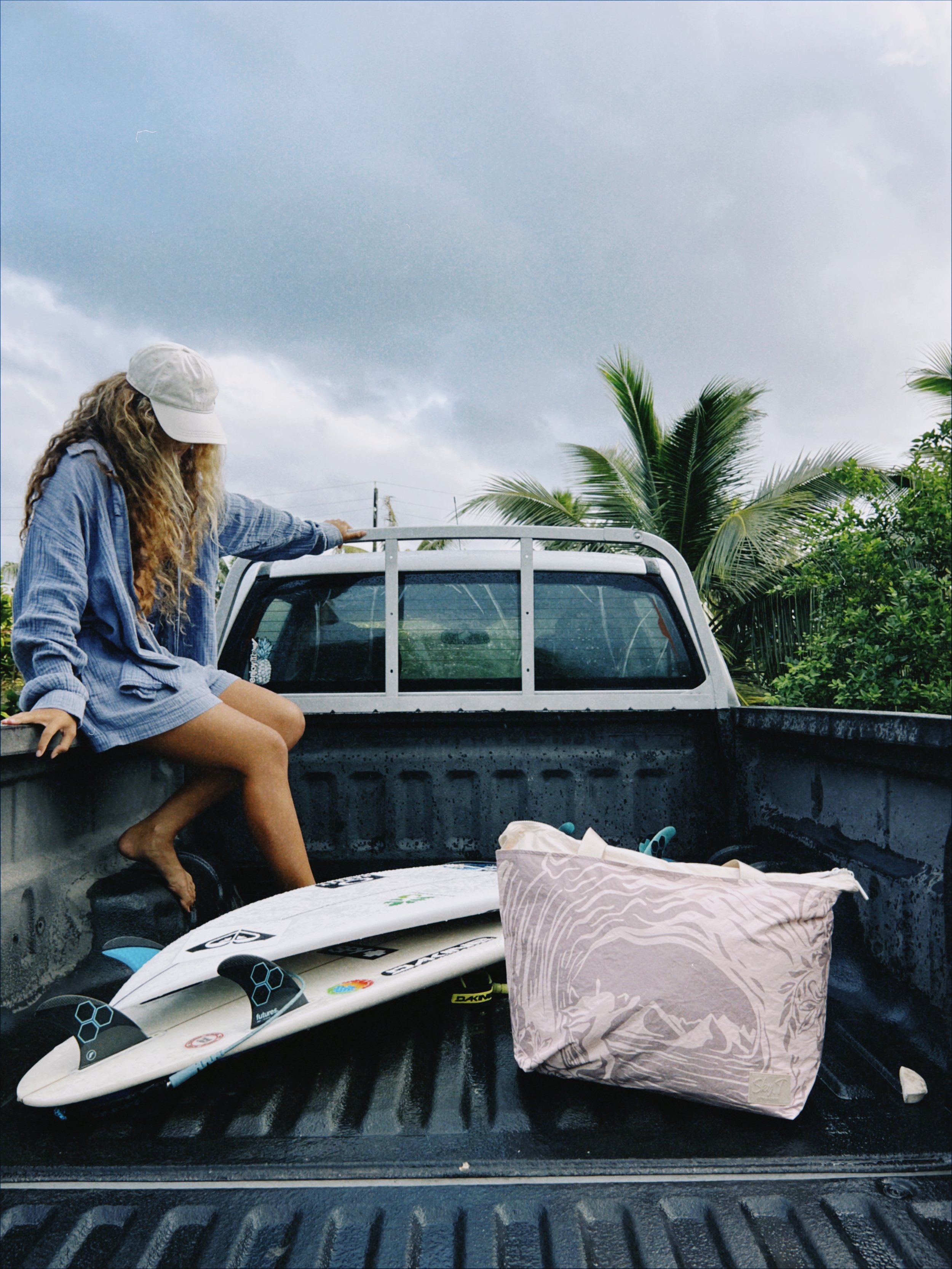 Une jeune femme assise dans la caisse d’un camion, avec une planche de surf, un sac et des vêtements, sous un ciel nuageux, entourée de palmiers.
