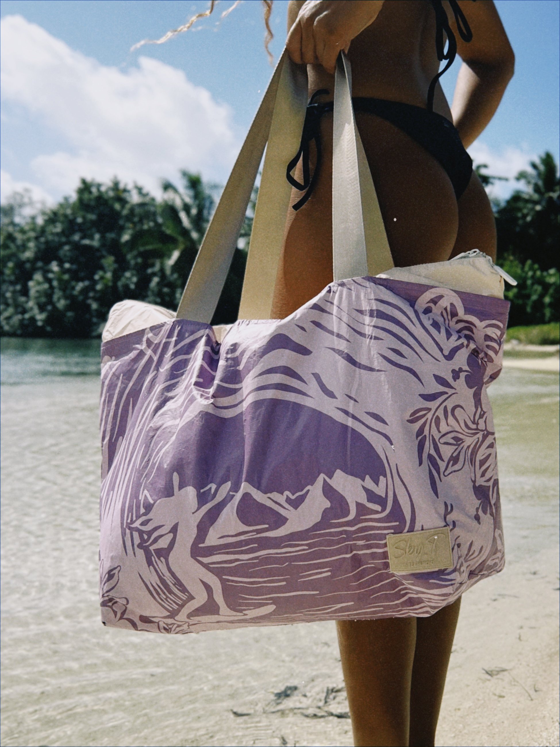 Personne vue de dos portant un grand sac de plage violet avec des illustrations de vagues et un arbre, sur une plage avec de l'eau et des arbres en arrière-plan.