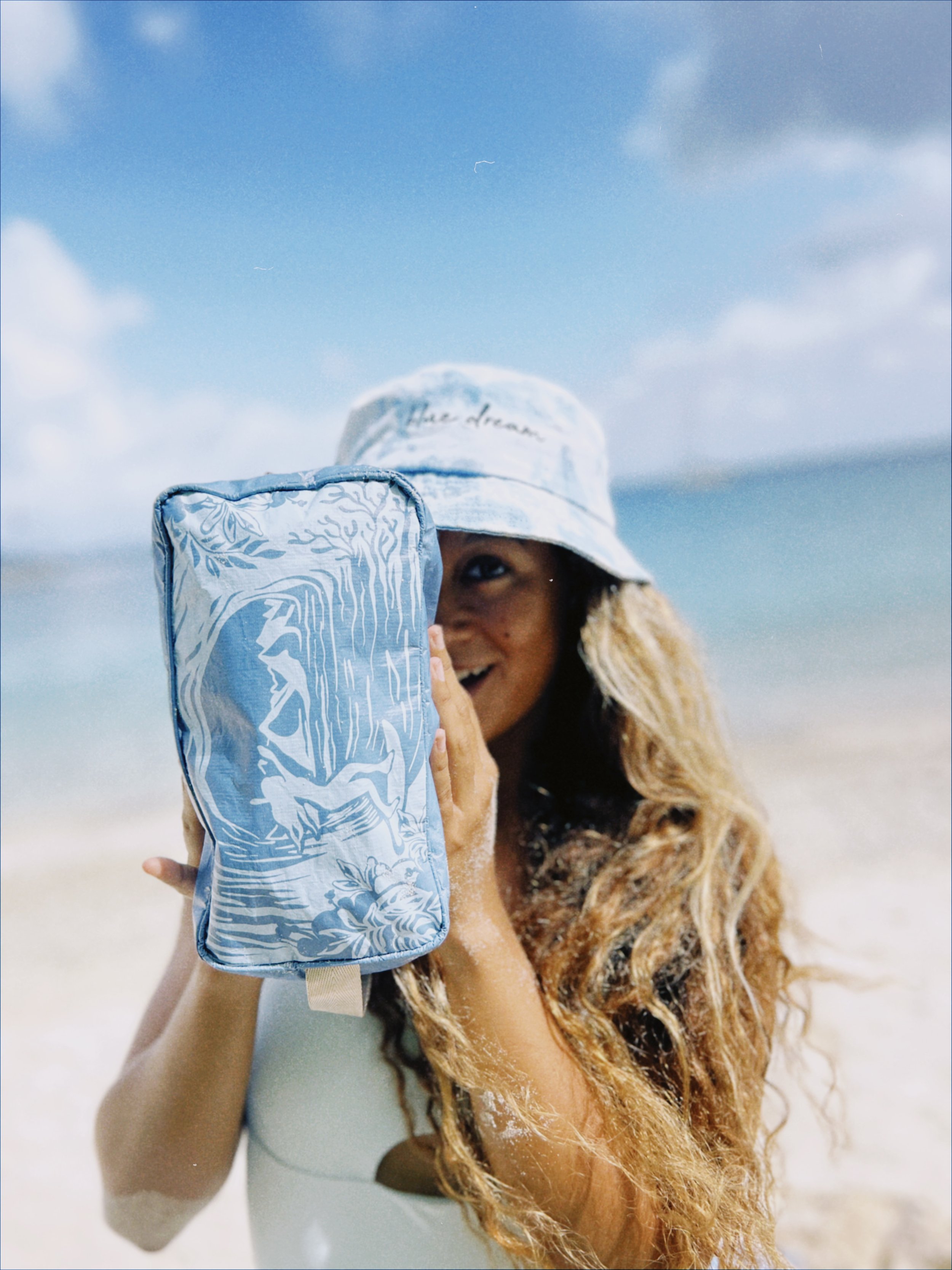 Une femme souriante avec de longs cheveux bouclés portant un chapeau à la plage, tenant une pochette ou un sac de plage bleu avec des motifs blancs.