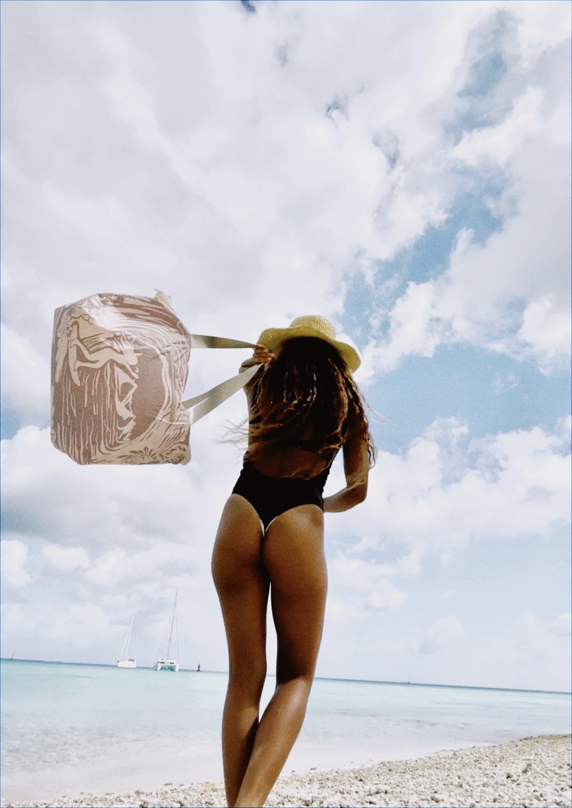 Femme portant un chapeau de paille, en maillot de bain noir, avec un grand sac, se tenant sur la plage face à la mer avec des bateaux en arrière-plan, sous un ciel nuageux.