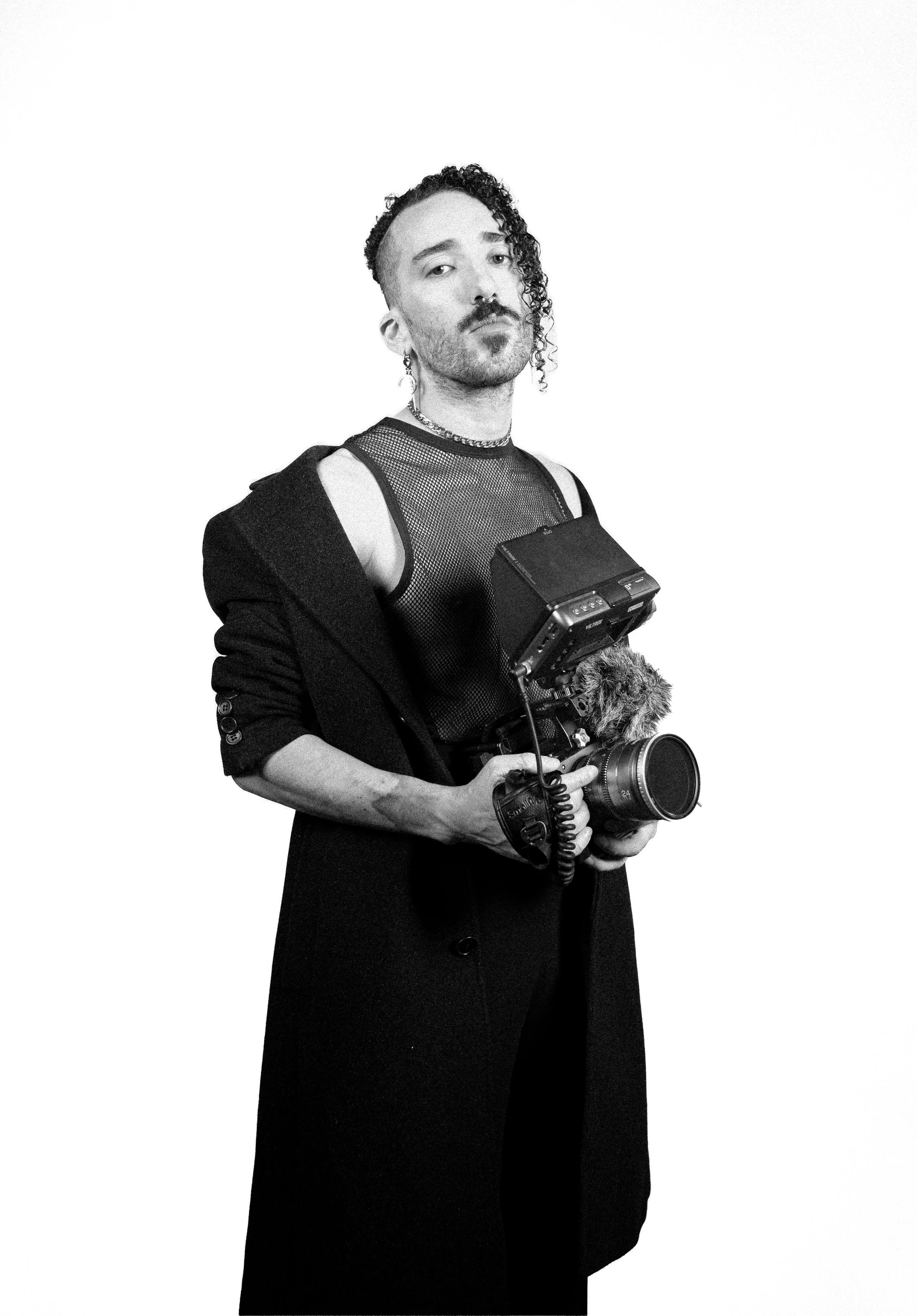 A person holding professional video recording equipment, wearing a mesh top, jacket, and jewelry, with a hairstyle featuring one side shaved and the other side curly.