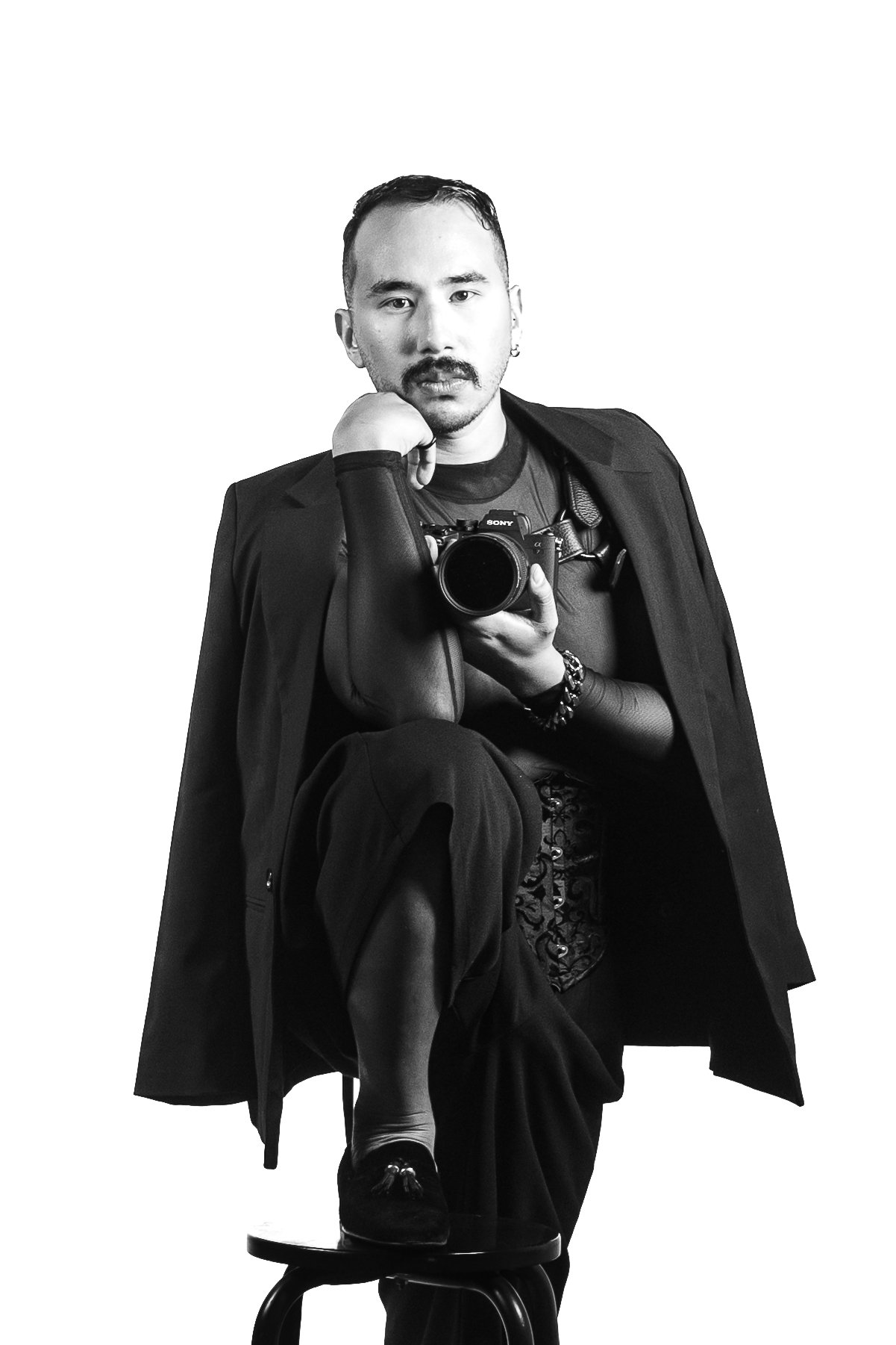 A person taking a selfie in front of a mirror, holding a camera, and posing with one knee on a stool, dressed in dark clothing and accessories.