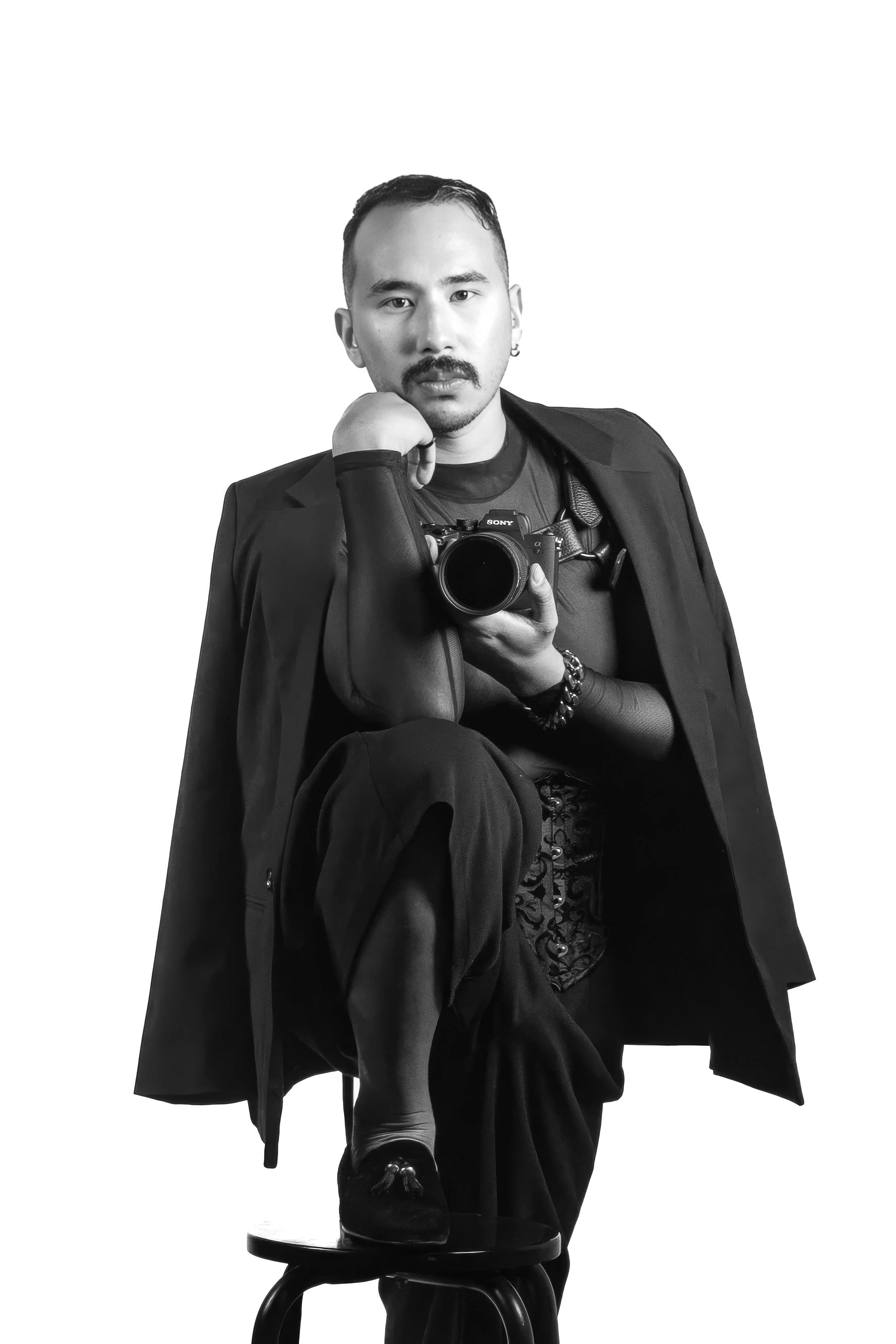 A person taking a black and white mirror selfie with a DSLR camera, sitting on a stool with one leg crossed over the other, dressed in fashionable clothing and accessories
