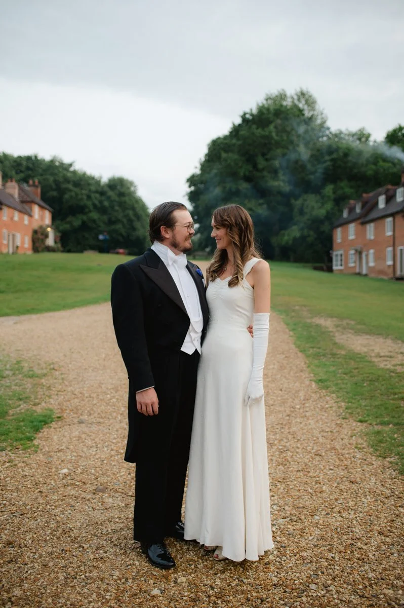 A groom and bride in wedding attire smiling and looking at each other on a gravel pathway outdoors with green trees and residential buildings in the background.