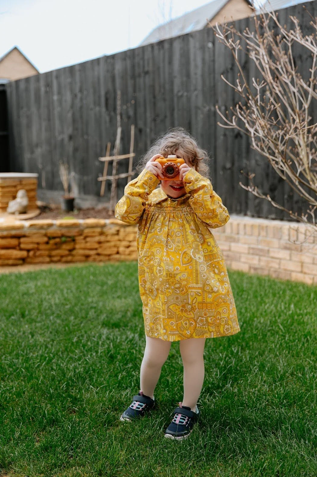 A young girl in a yellow patterned dress holding a camera, standing on grass in a backyard, with a wooden fence and leafless trees in the background.