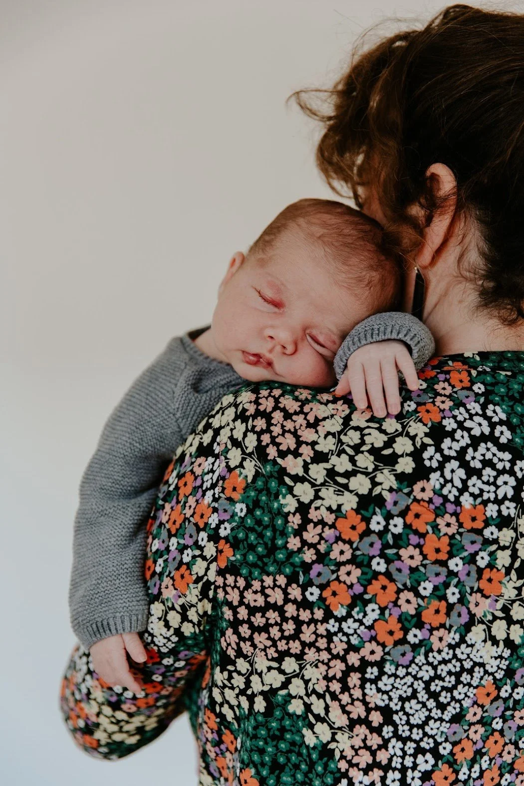 A baby sleeping on an adult's shoulder, with the adult wearing a floral shirt and holding the baby with one hand.