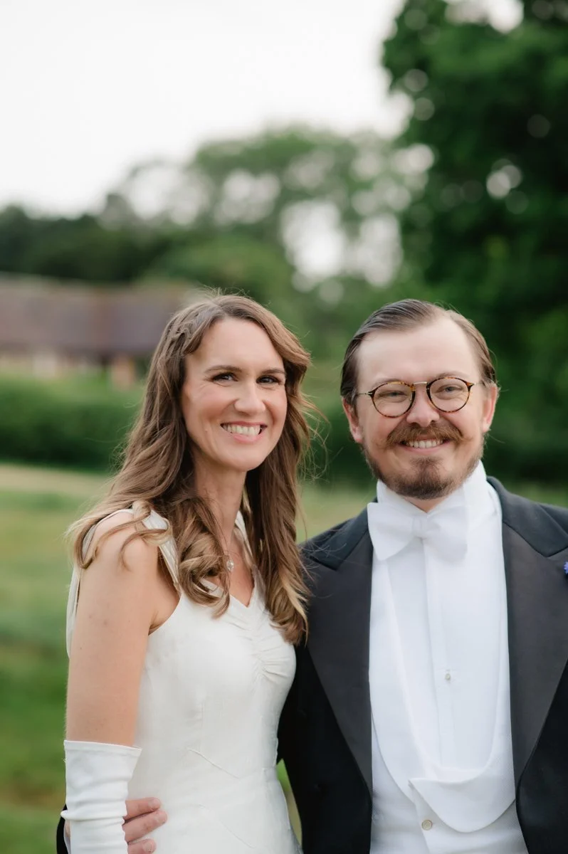 A smiling woman in a white formal dress with long, wavy hair standing next to a smiling man in formal attire, including glasses, a white bow tie, and a black tuxedo, outdoors with green trees in the background.