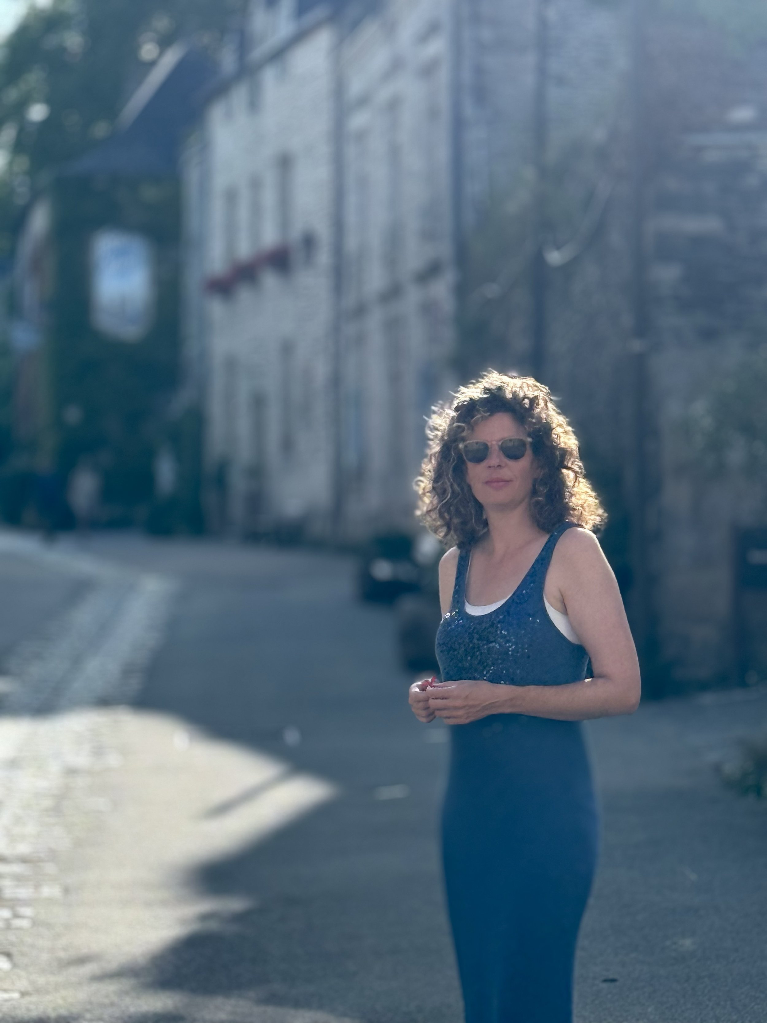 Woman with curly hair and sunglasses standing on a city street, wearing a sparkly blue tank top and dark pants, with a building and cars in the background.