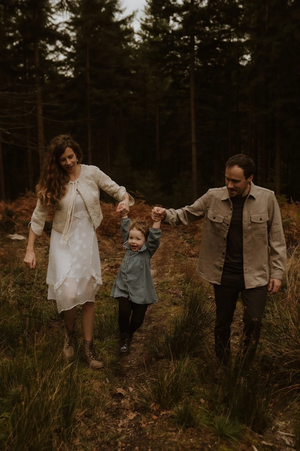 A family of three walking on a forest trail, holding hands, smiling, and enjoying nature during the daytime.