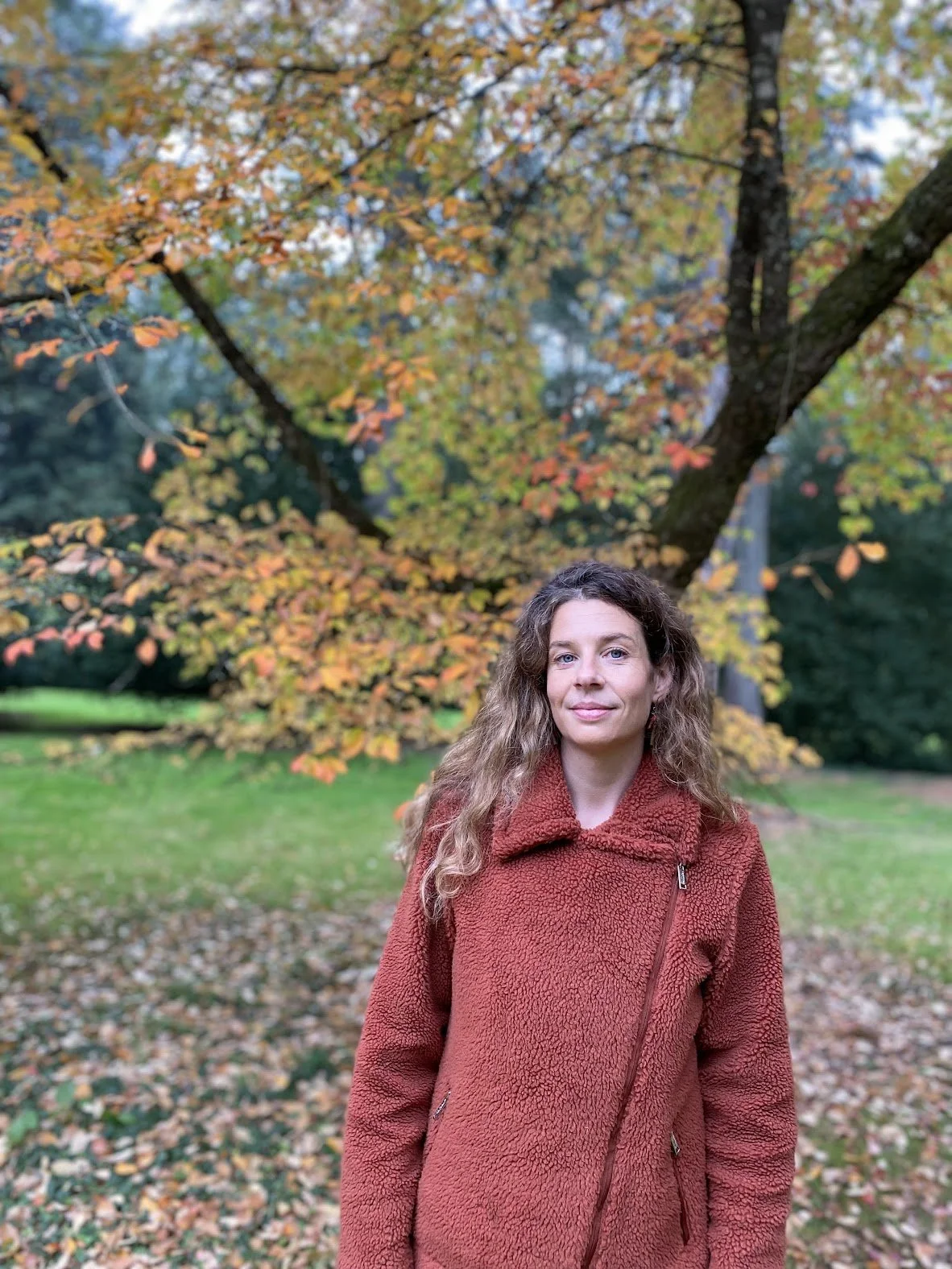 A woman with long curly hair wearing a rust-colored coat standing outdoors in front of an autumn tree with orange and yellow leaves.
