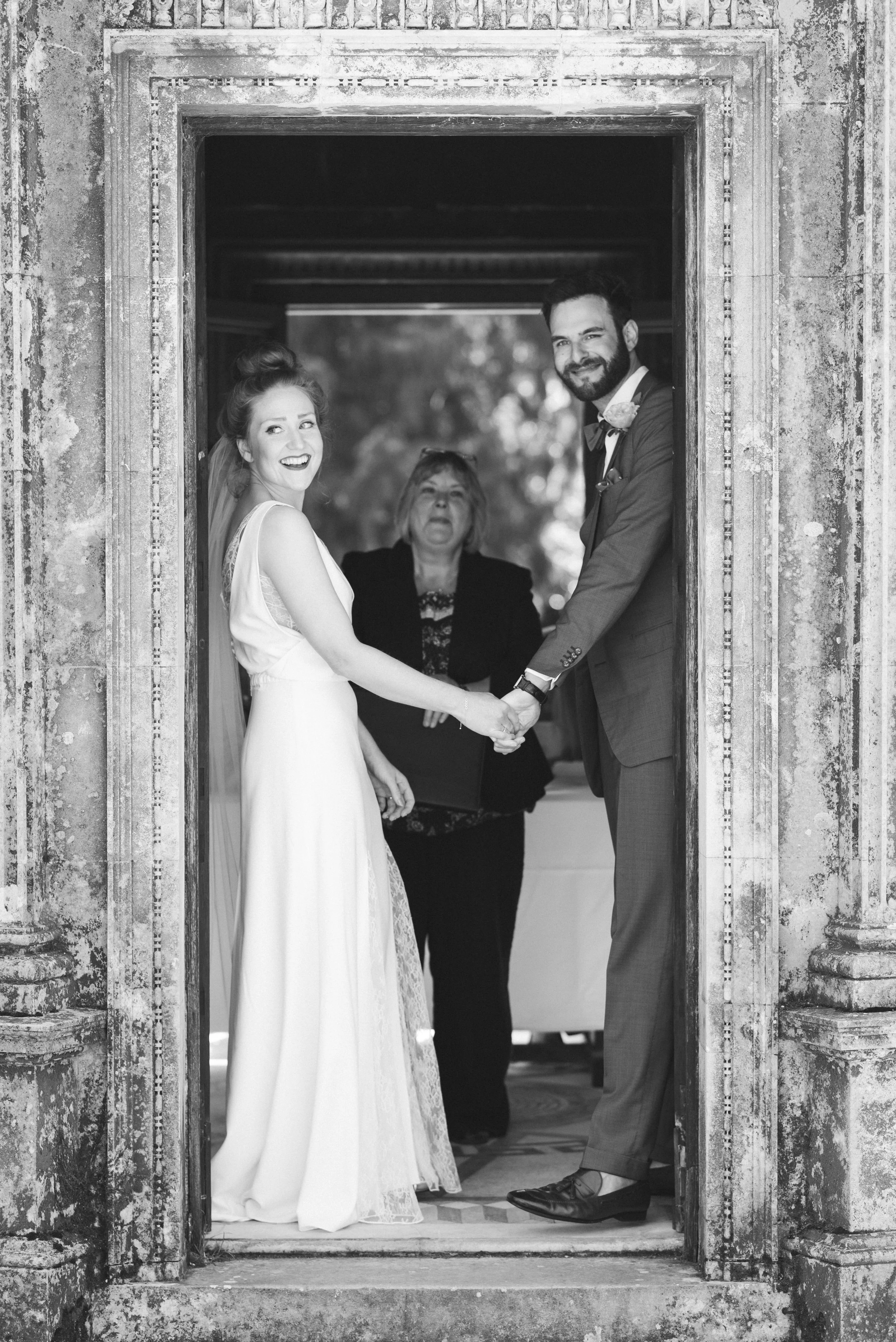 A black and white photo of a bride and groom holding hands and smiling in a doorway, with a woman in the background.