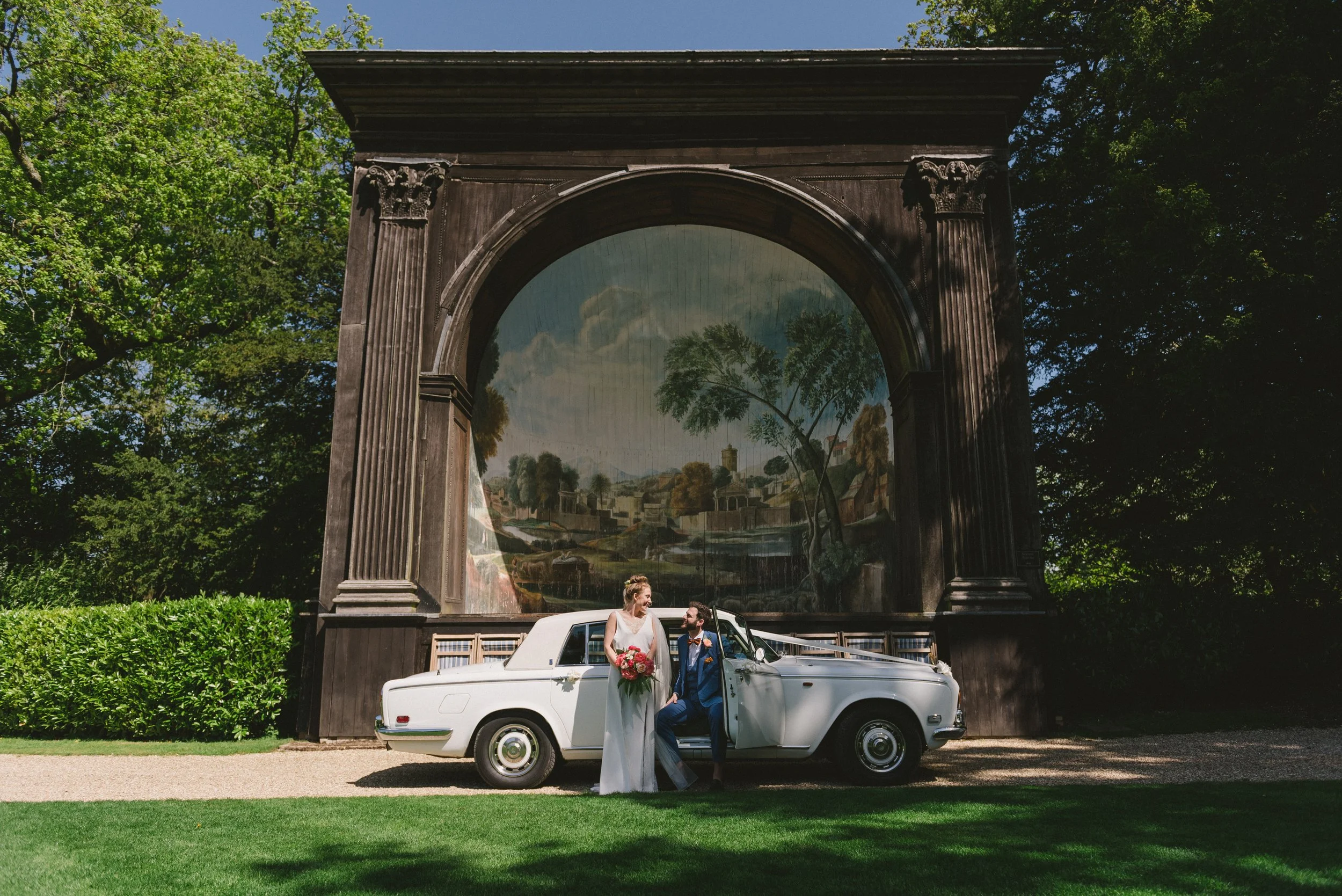 A bride and groom standing beside a white vintage car on a grassy area in front of a large decorative arch with a landscape painting. The bride is holding a bouquet of flowers, and they are looking at each other.