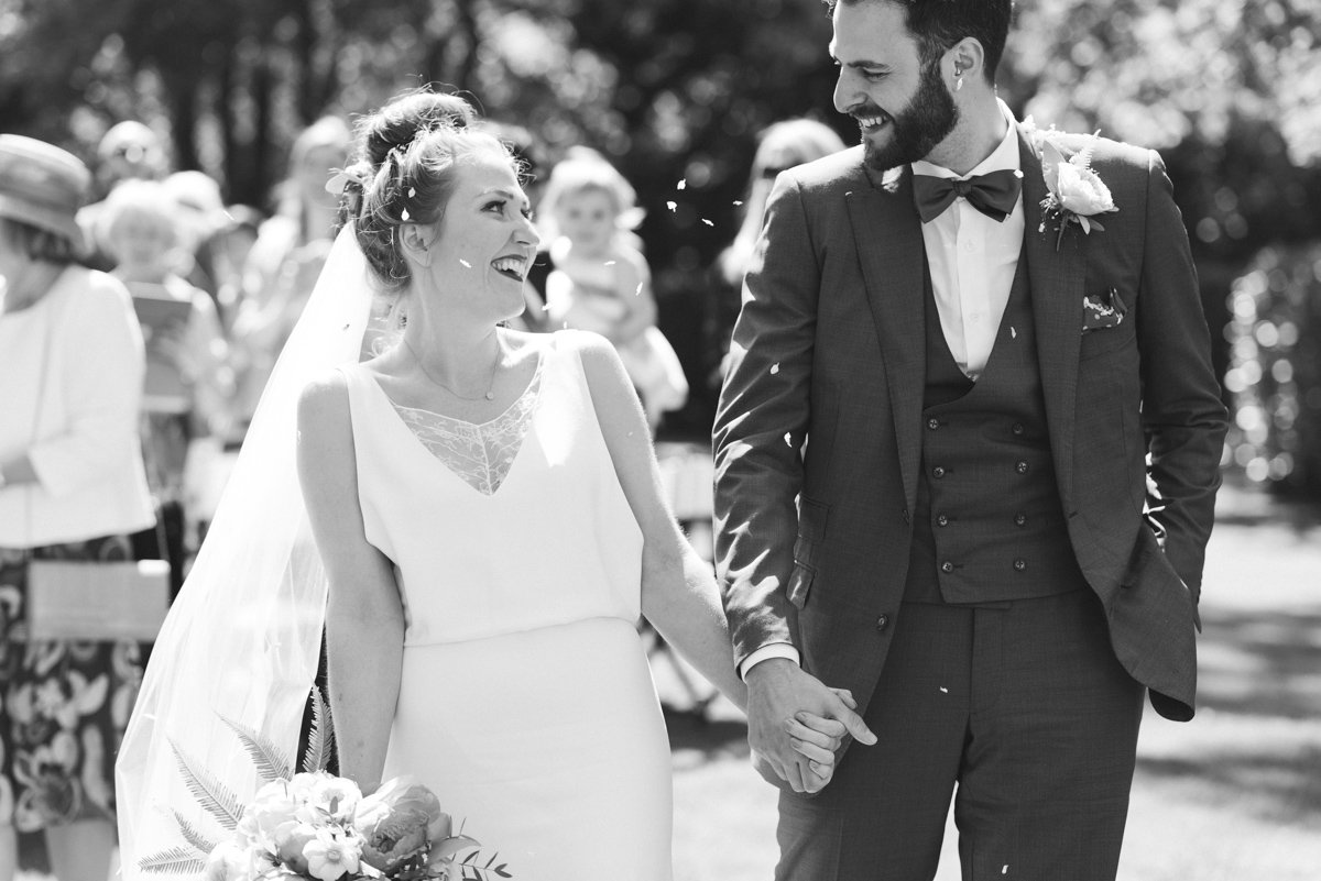 A bride and groom holding hands and smiling during their outdoor wedding ceremony, with guests in the background.