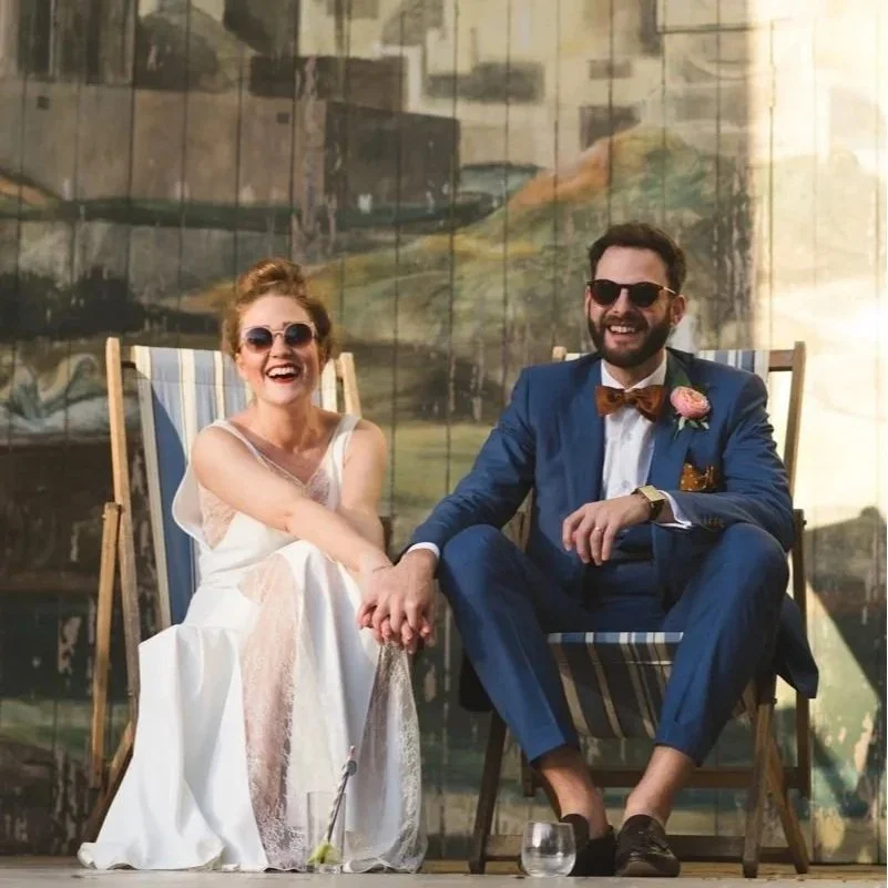 A smiling couple, dressed in formal wedding attire, sitting on deck chairs, holding hands, and wearing sunglasses.