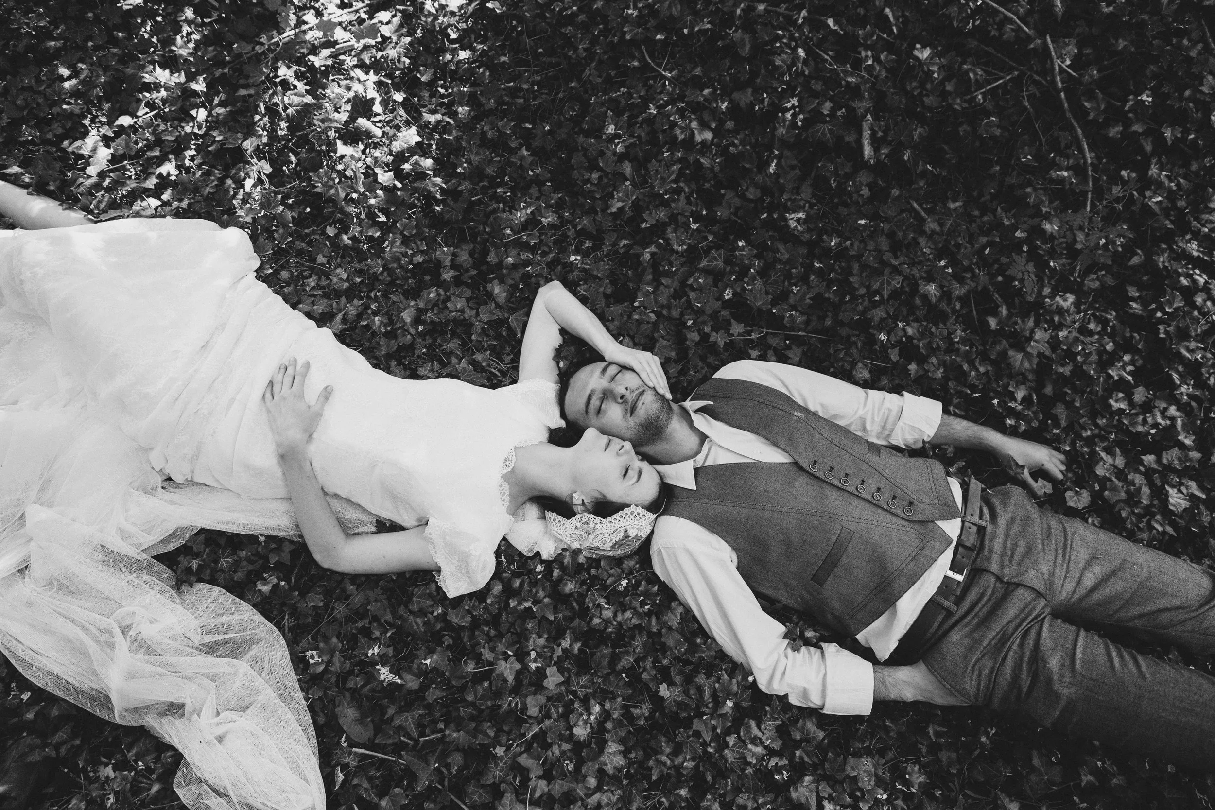 A bride and groom lie on a bed of ivy leaves, with the bride resting her head on the groom's shoulder and his eyes closed.