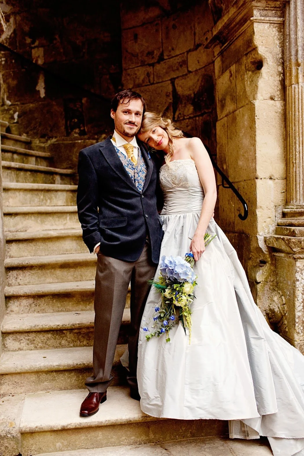 A wedding couple, man in a dark blazer, woman in a wedding dress, standing on stone steps inside a historic building with stone walls.