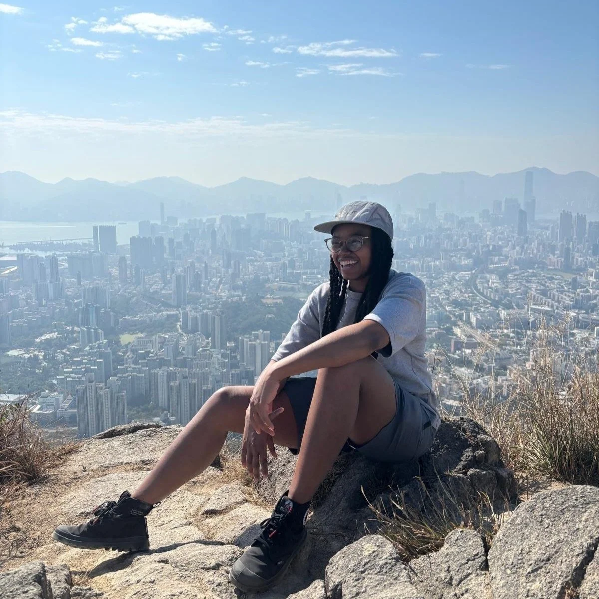 A person sitting on rocks at a mountain lookout with a city skyline, mountains, and blue sky in the background, smiling and wearing a hat, glasses, a gray shirt, shorts, and hiking shoes.