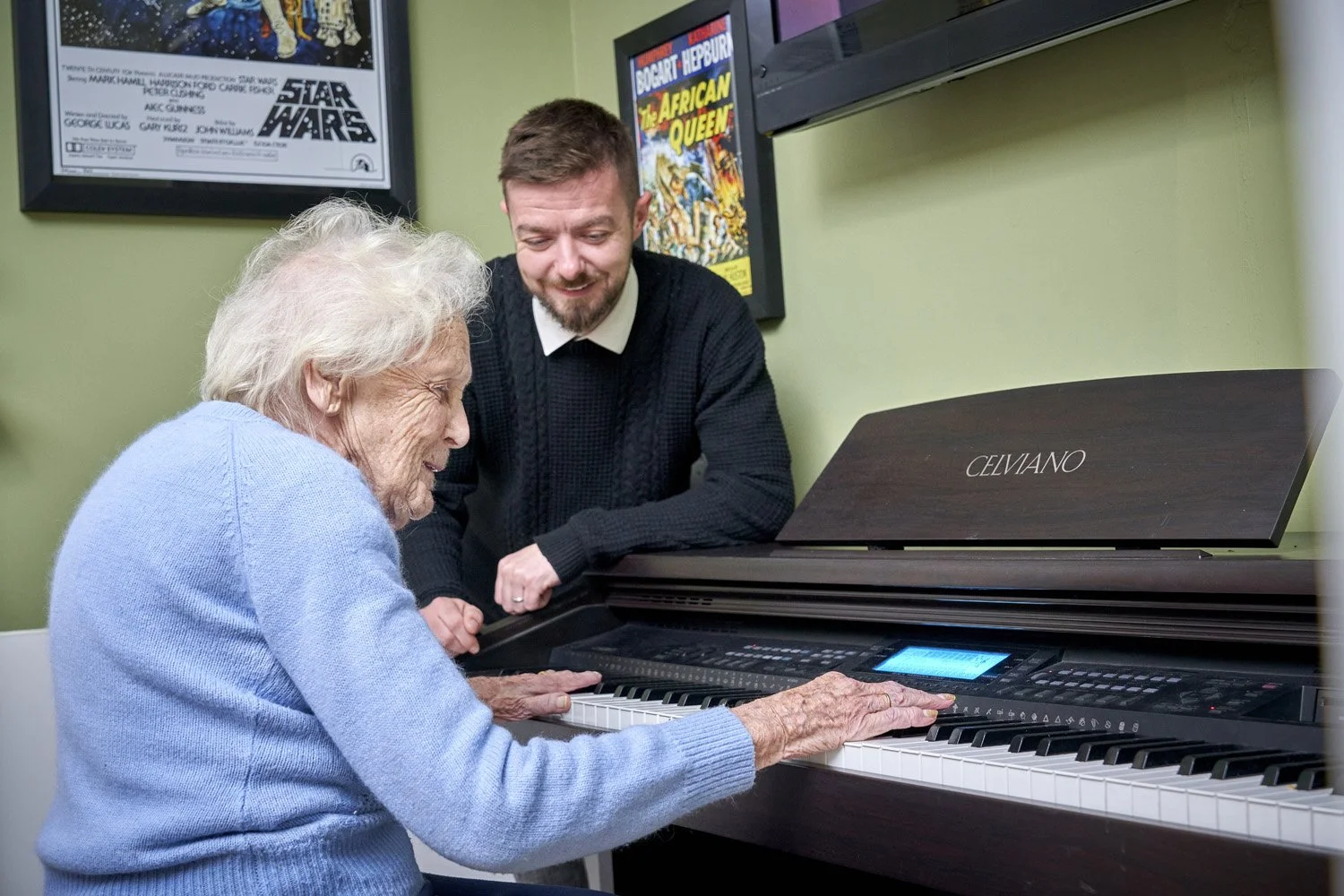 An elderly woman and a middle-aged man are sitting beside a digital piano, with the woman playing and the man leaning in, smiling. The room has framed posters on the wall, including one for Star Wars and another for African Queen.