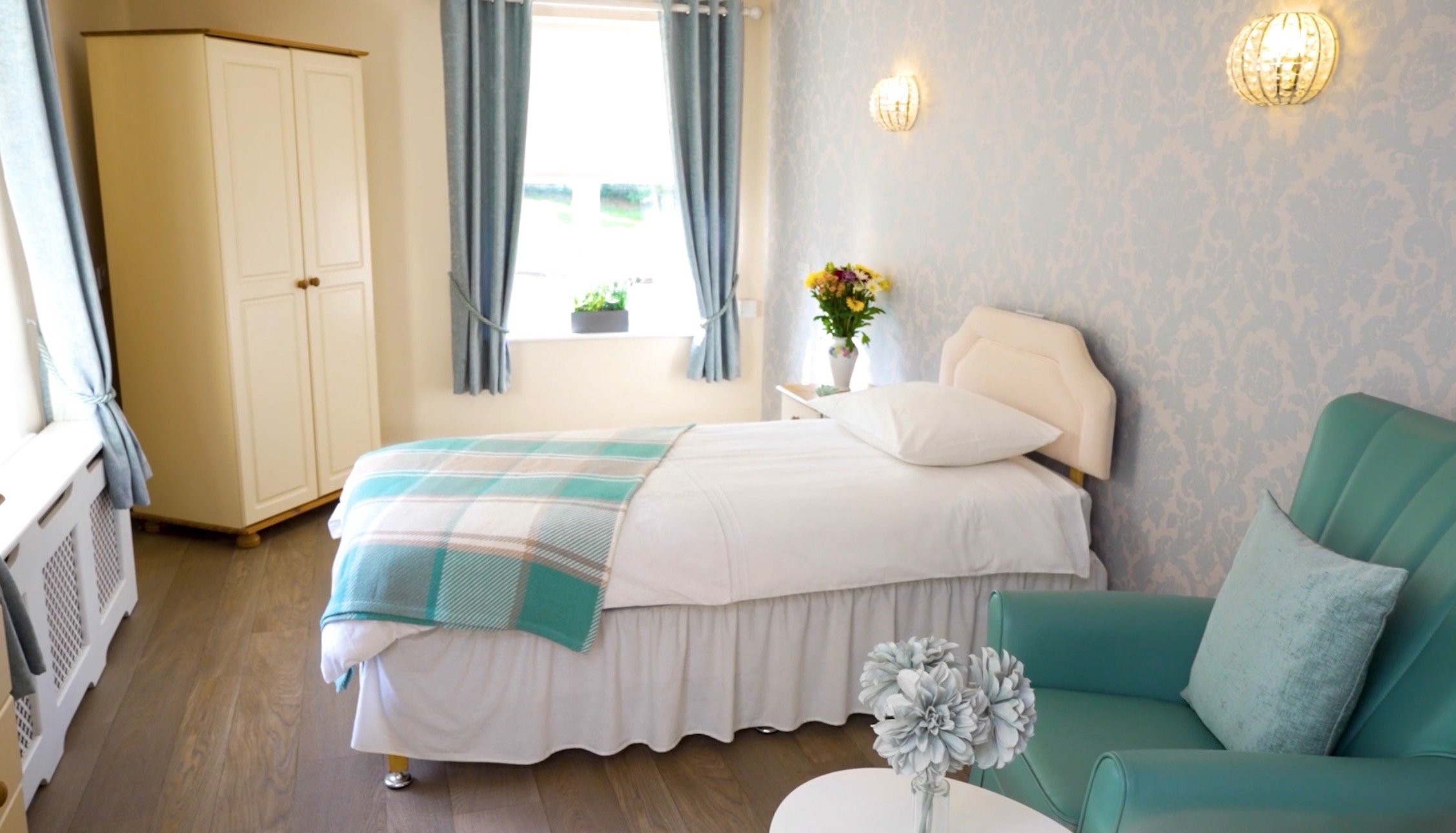 A cozy bedroom with a single bed, blue and white color scheme, and a window with blue curtains. There is a green armchair with pillows, a white bedside table with flowers, and a wardrobe.
