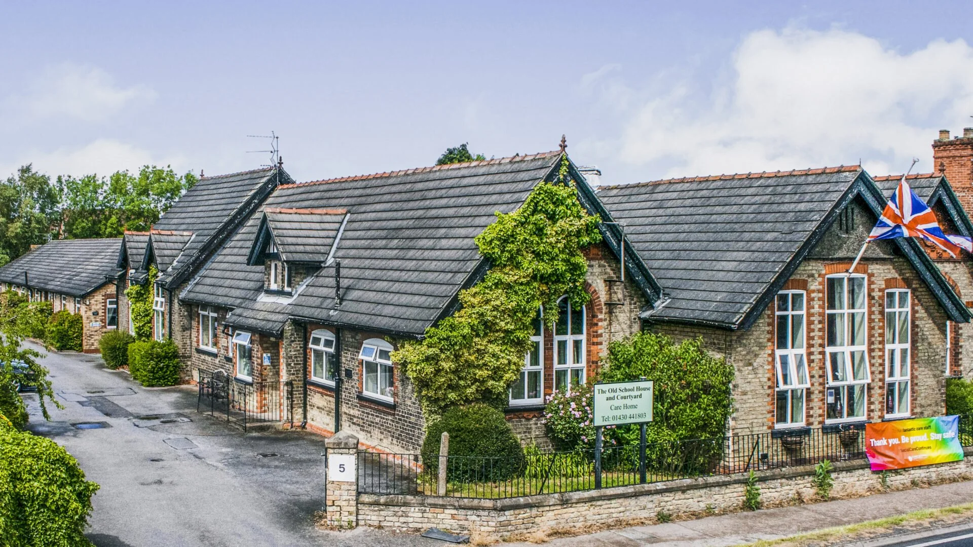 A row of old brick houses with slate roofs, green plants and bushes, a Union Jack flag, and a colorful sign, under a partly cloudy sky.