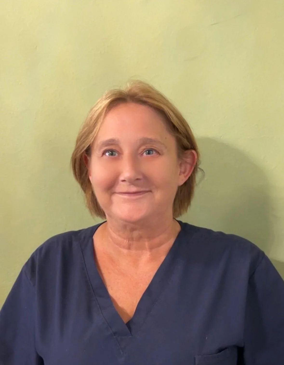 Woman in navy medical scrubs smiling in front of a light green wall.