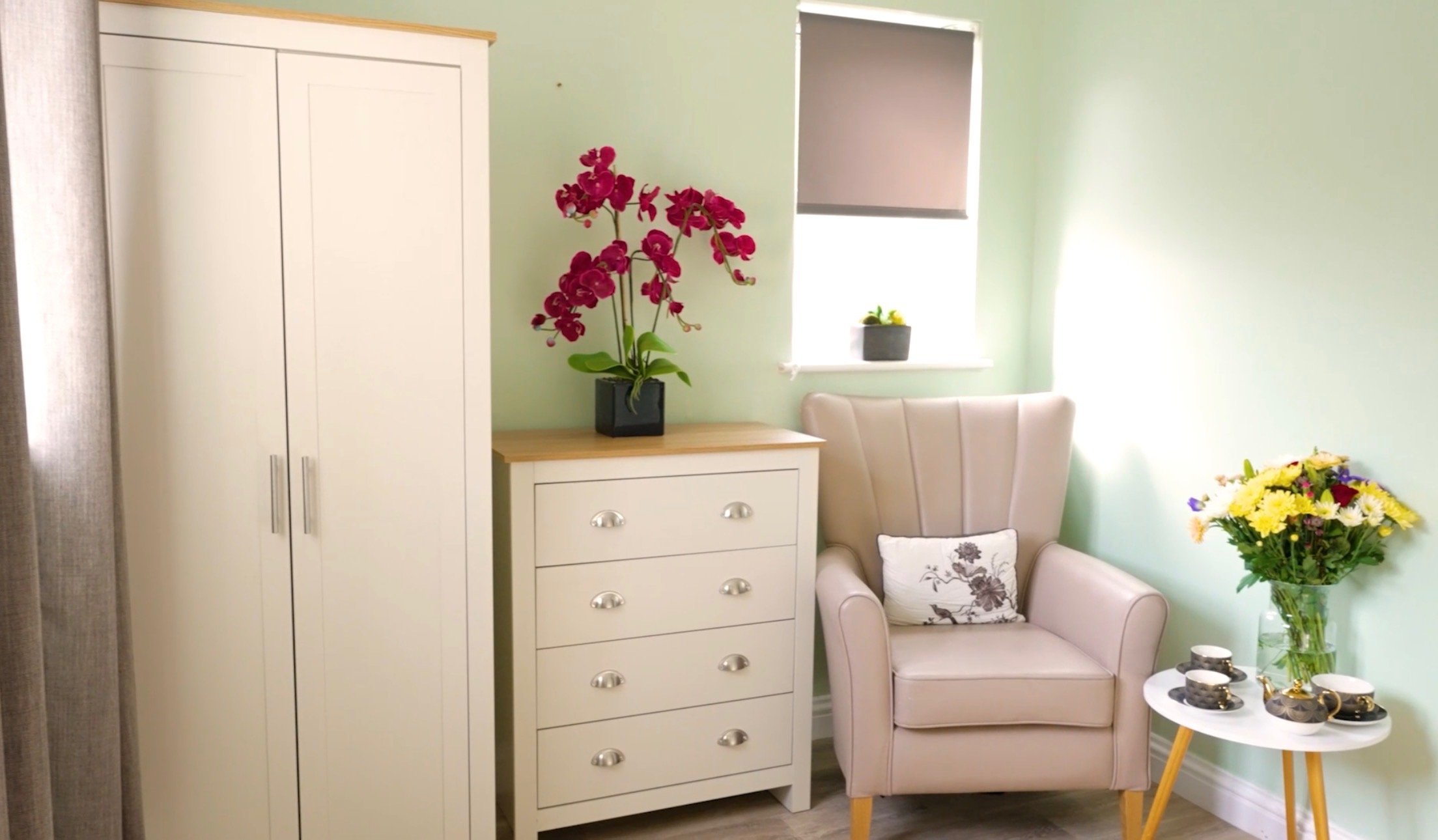 A cozy corner with a white wardrobe, a white dresser topped with a pink orchid, a beige armchair with a floral pillow, a window with a rolled-up gray blind, and a small round table with a vase of colorful flowers and tea cups.