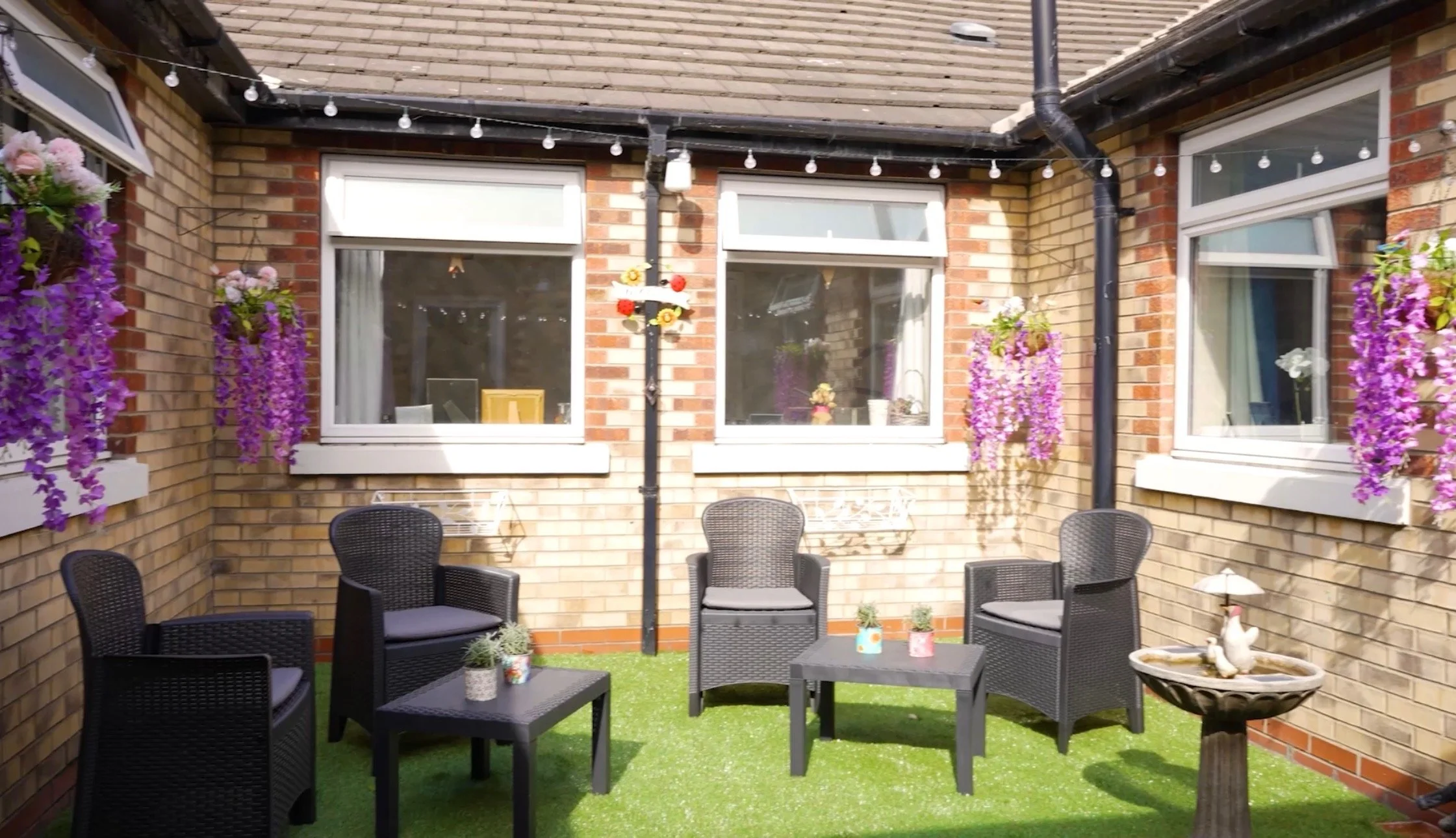 A small outdoor patio area with artificial grass, black wicker furniture including chairs and a coffee table, potted plants, hanging purple flowers, and a bird bath with a decorative duck. The patio is enclosed by brick walls with three windows and s