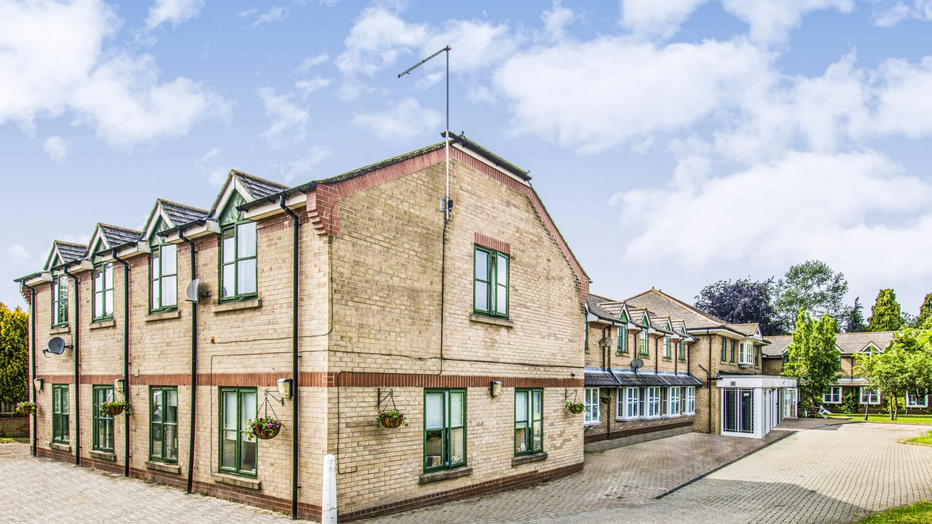 A multi-story residential building with brick walls, green window frames, and hanging flower baskets, situated in a landscaped area with trees and a paved pathway under a partly cloudy sky.