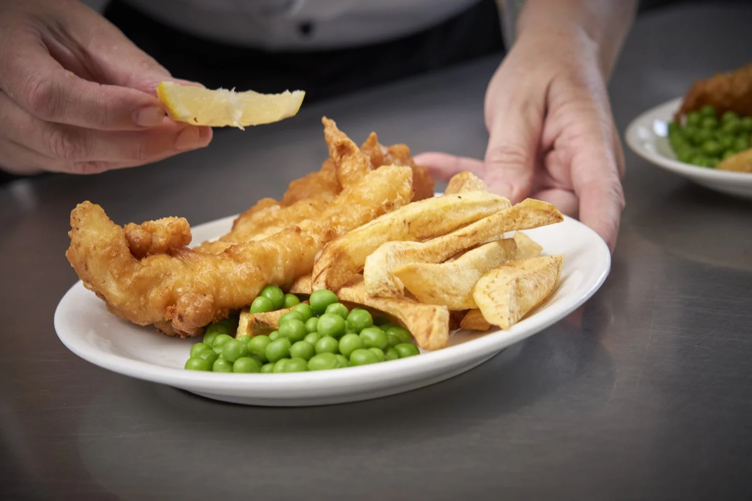 A white oval plate with fried fish, potato wedges, green peas, and French fries, with a lemon wedge being added.