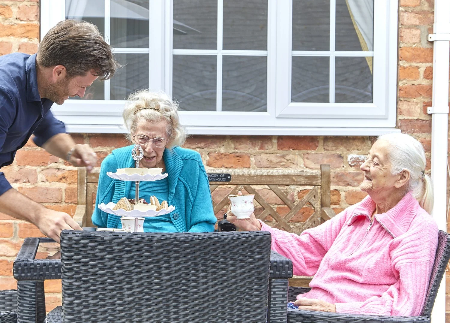 A man serving tea to two elderly women garden seating outdoors, with a cake on a stand and a brick wall background.
