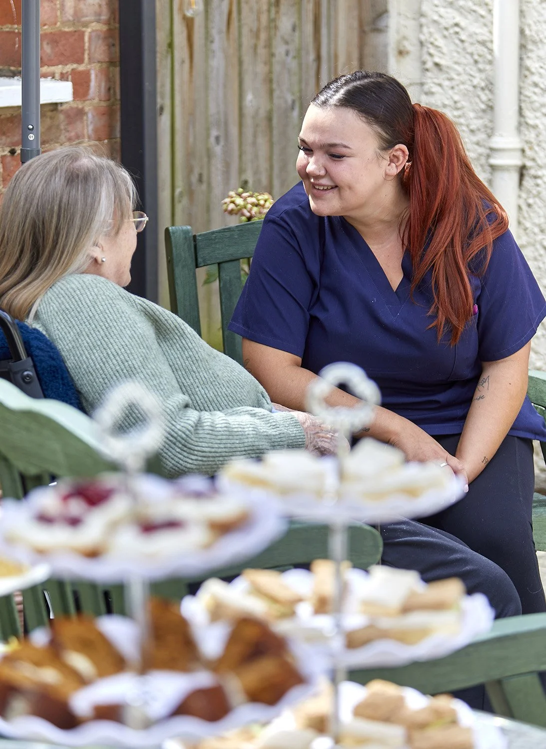 A young woman in a navy scrubs talking with an elderly woman sitting on a green bench, with a brick and wooden fence in the background. A blurry tiered tray of assorted desserts is in the foreground.