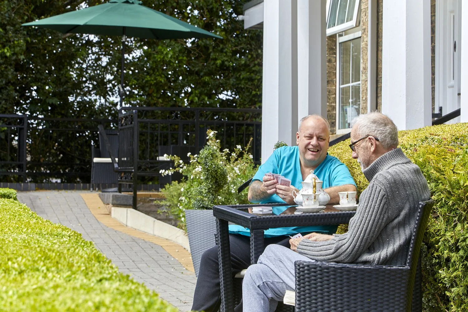 Two men sitting outdoors at a table playing cards, with a teapot, cups, and a sugar bowl, in a garden patio with green bushes, a brick ramp, and an umbrella in the background.