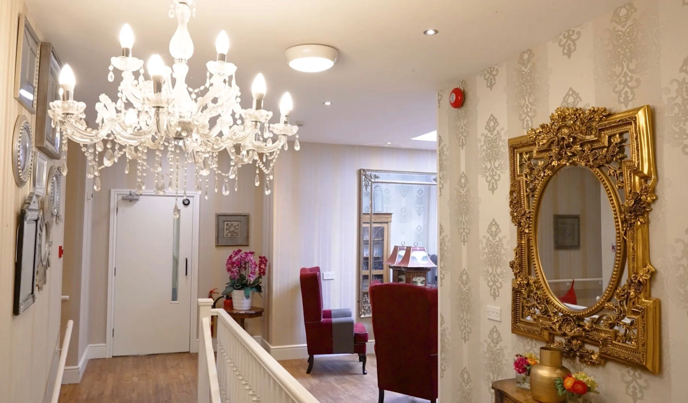 Interior of a well-decorated room featuring a large crystal chandelier, gold-framed mirror, red chairs, patterned wallpaper, and floral decorations.