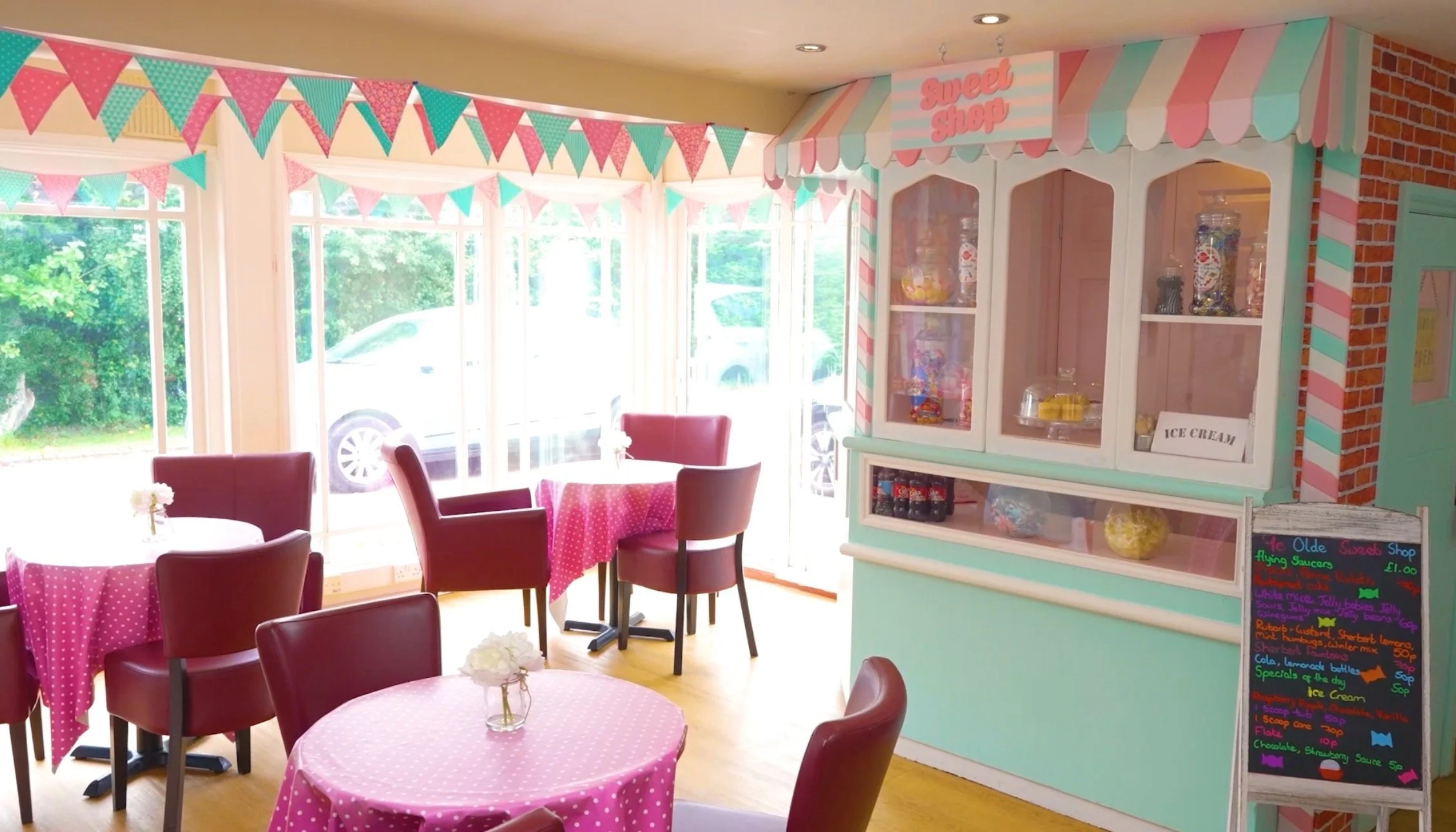 Interior of an ice cream shop with pink and teal decorations, pink polka dot tablecloths, maroon chairs, a colorful candy display, and a chalkboard menu.