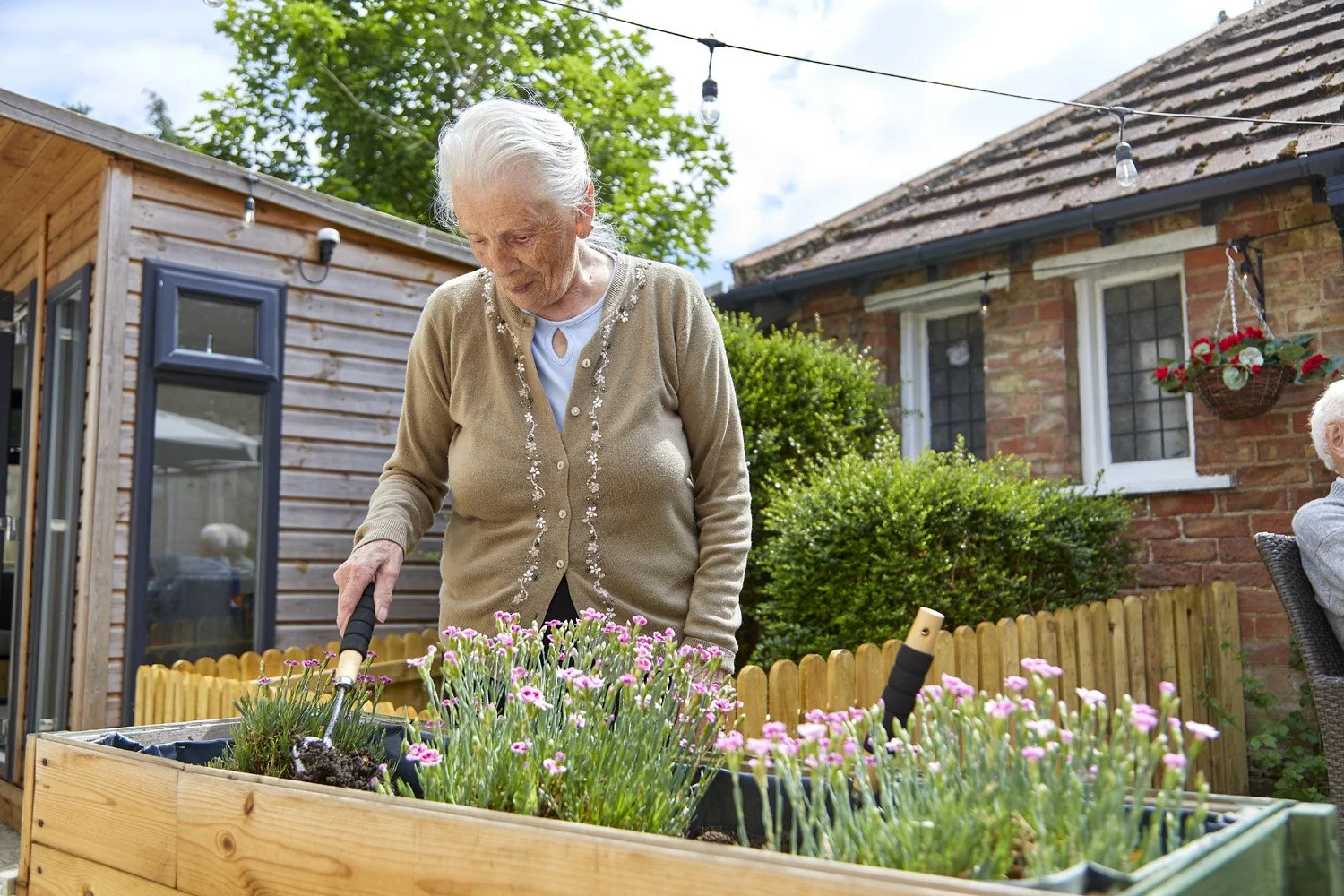 An elderly woman tending to pink flowers in a raised garden bed in her backyard, with a wooden shed, brick house, and a woman sitting on a chair in the background.