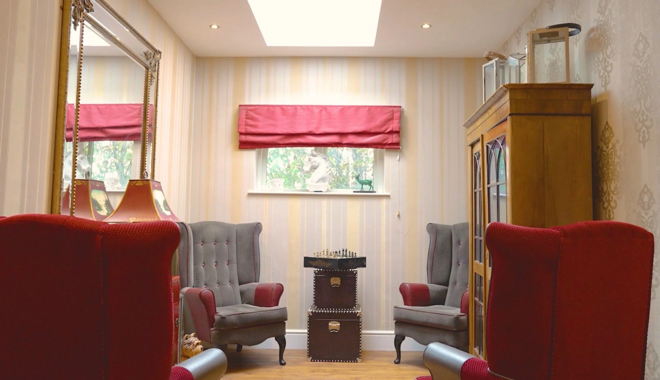 A cozy sitting room with two gray armchairs facing each other, two red velvet chairs, a small chessboard table, a tall wooden cabinet, mirrors, and a window with a pink valance and decorative items on the sill.