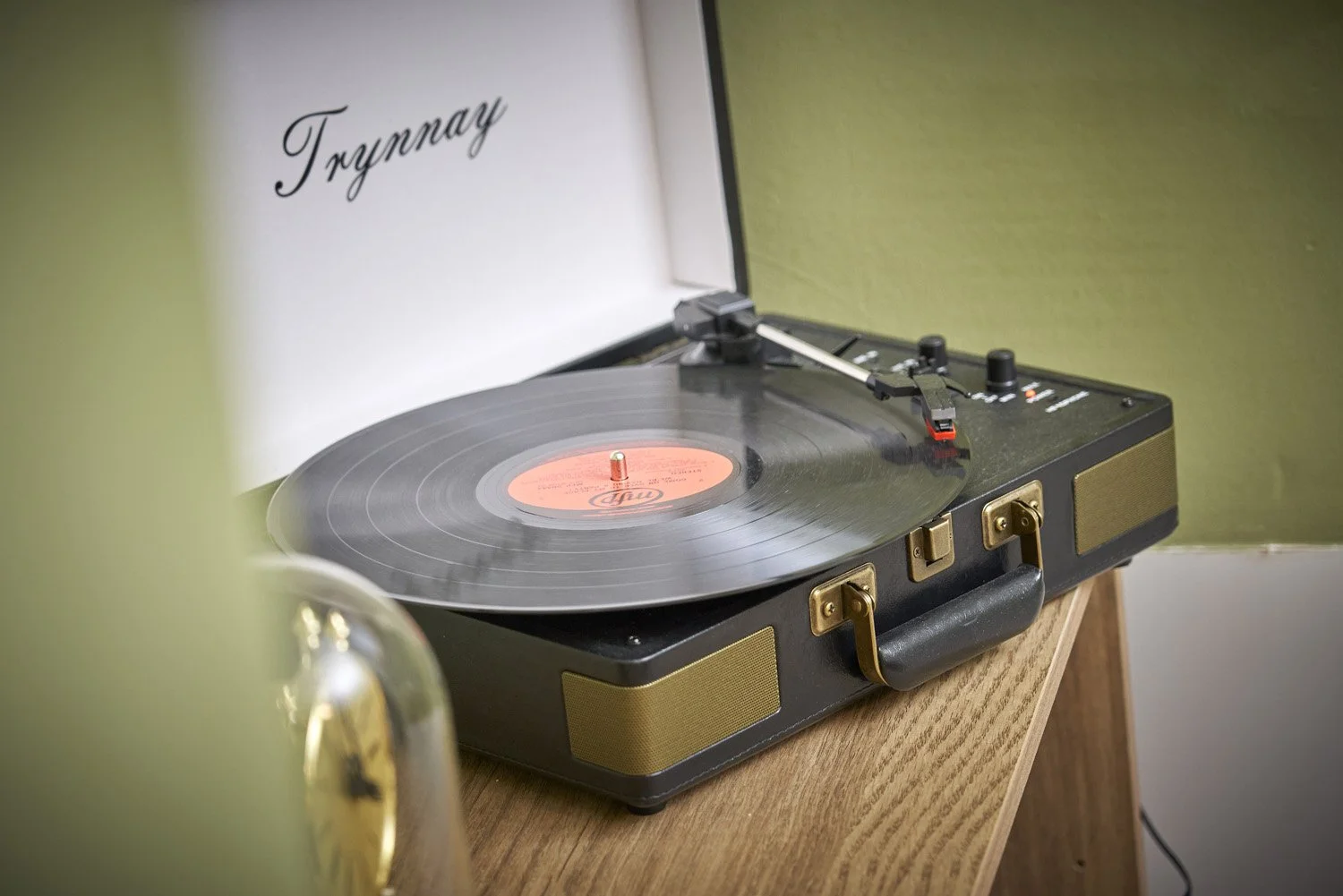 A vintage portable record player playing a vinyl record, placed on a wooden surface with a green and beige background, and a partial view of a metallic object in the foreground.