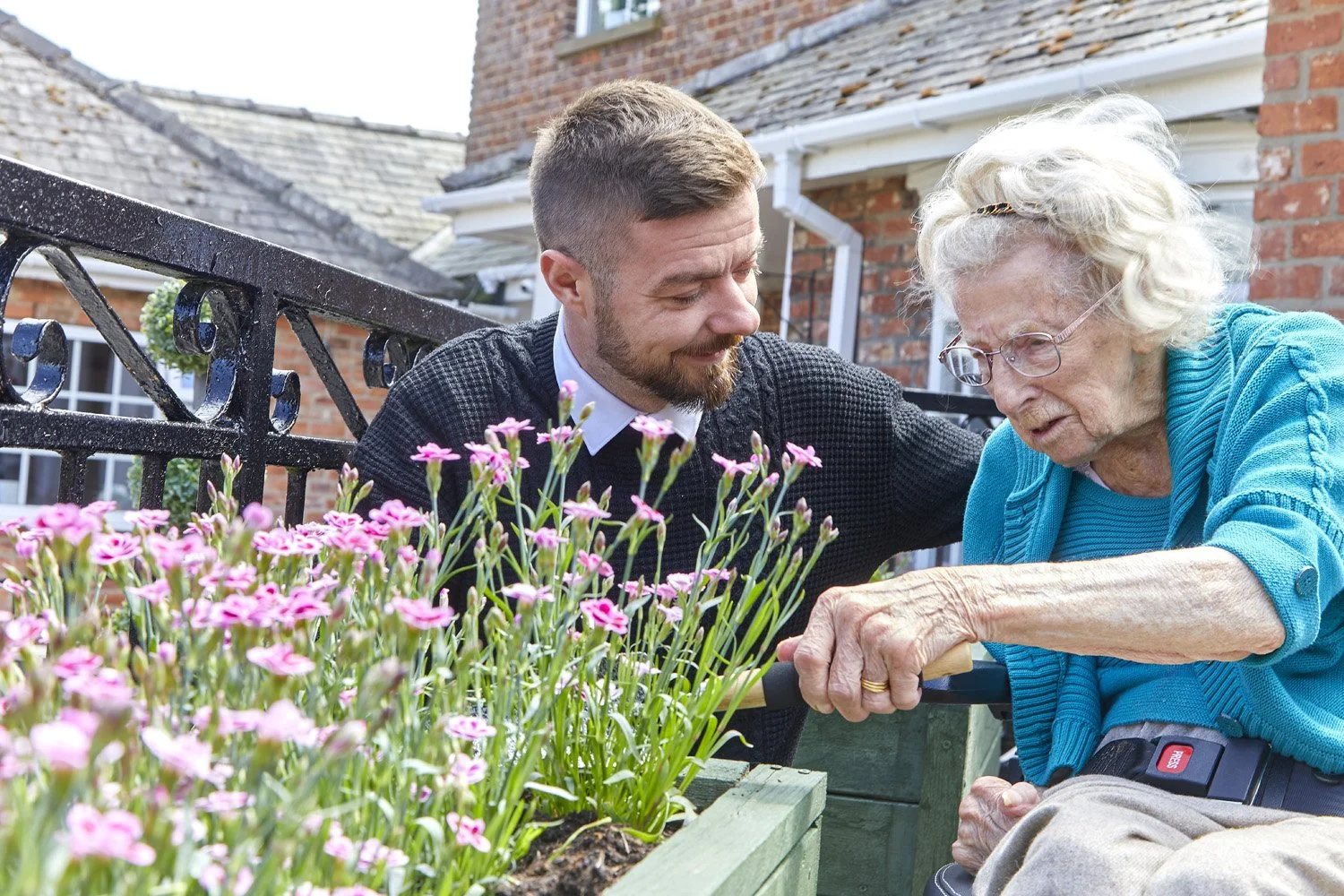A young man helps an elderly woman in a wheelchair tend to pink flowers in a garden on a sunny day, with brick buildings in the background.