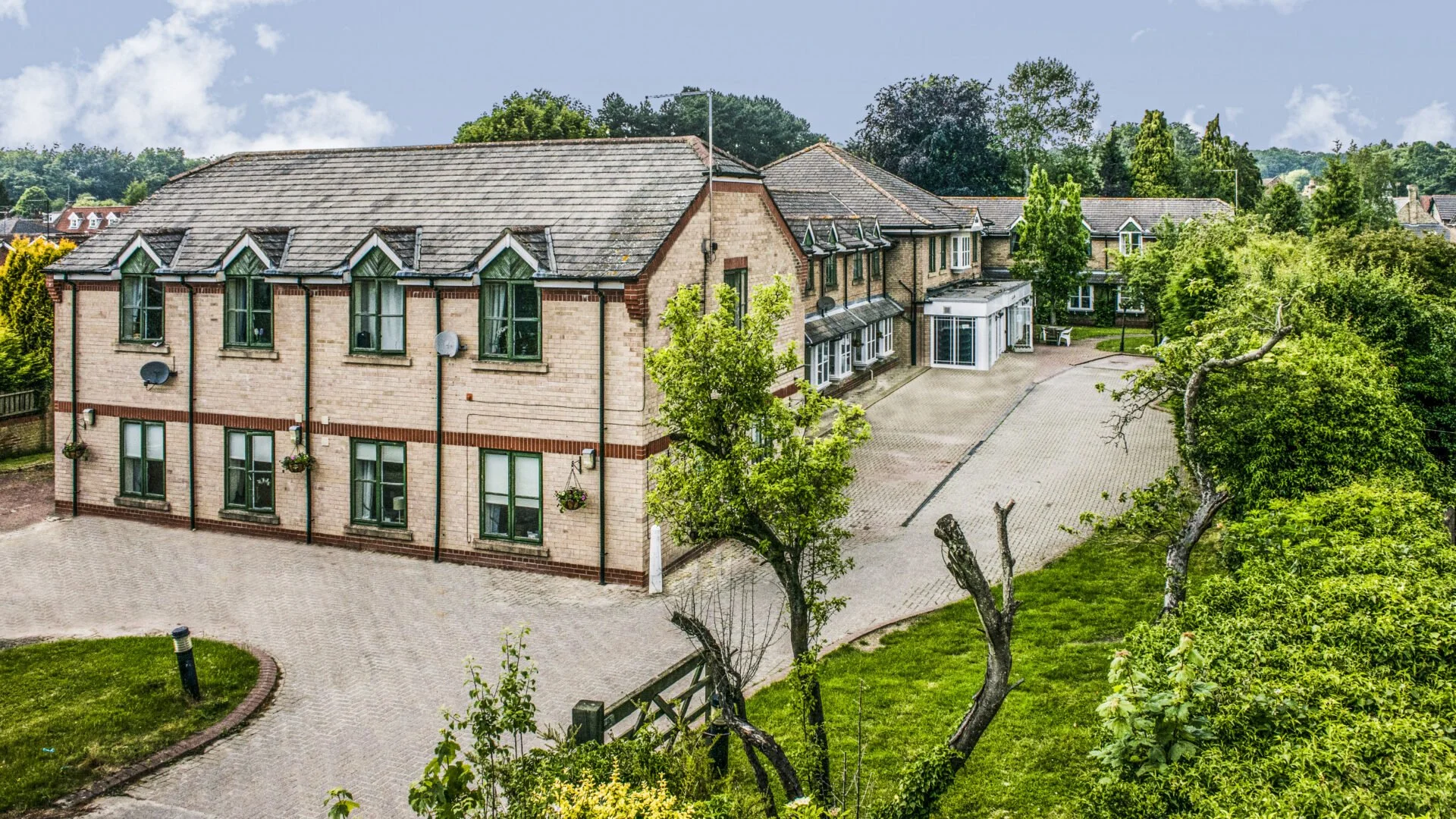 A large brick building with multiple windows and a gray tiled roof, surrounded by green trees and bushes, with a paved driveway and a grassy area.